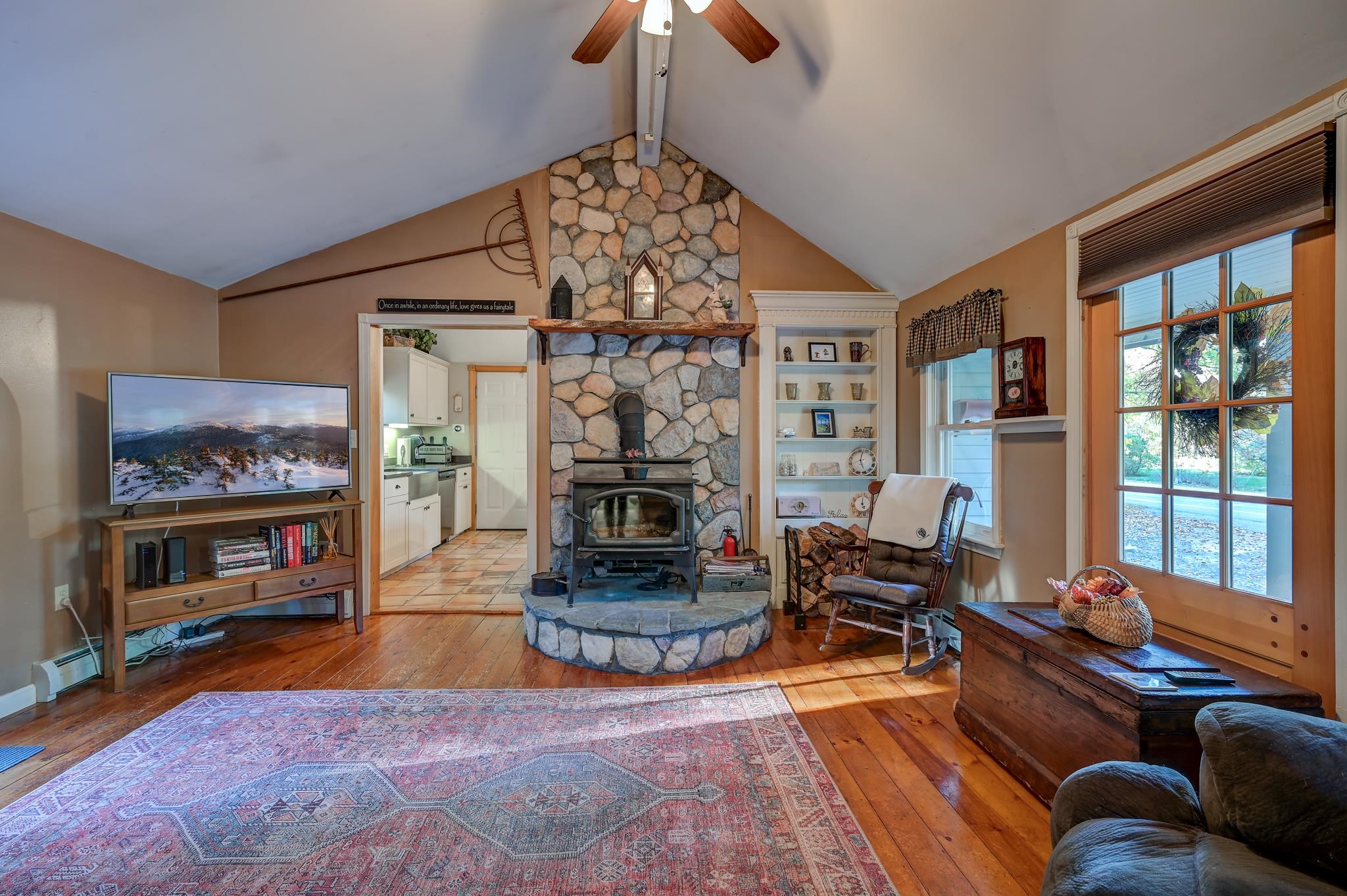Living room with a tall stone fireplace, wood stove, vaulted ceiling, and large sunlit windows.