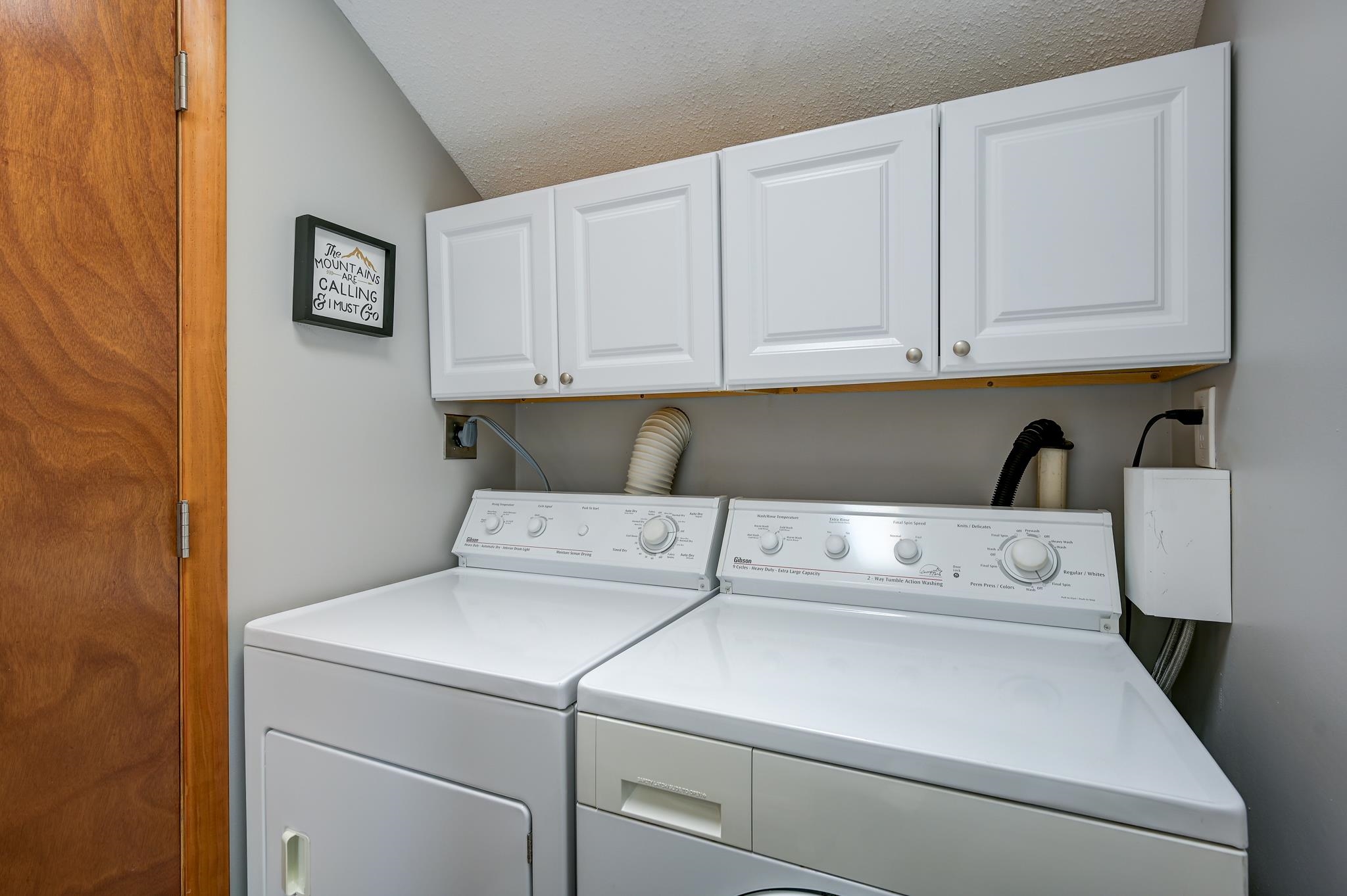 Laundry area with a washer and dryer beneath white cabinets and a framed mountain-themed sign.