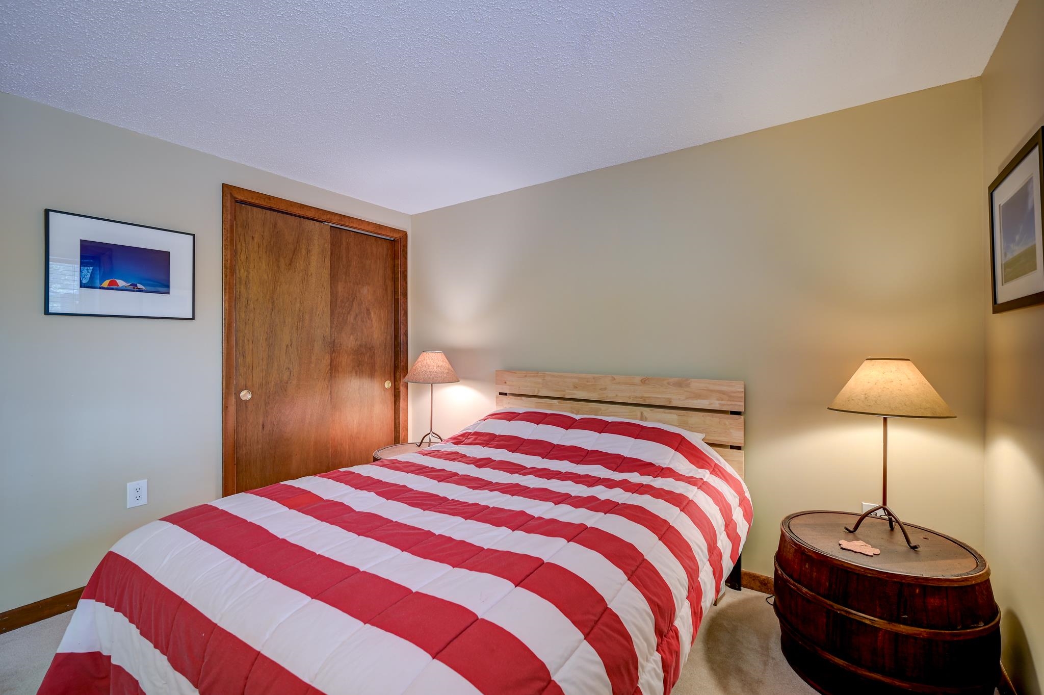 Bedroom with a red-and-white striped quilt, wood closet door, and barrel-shaped bedside table.