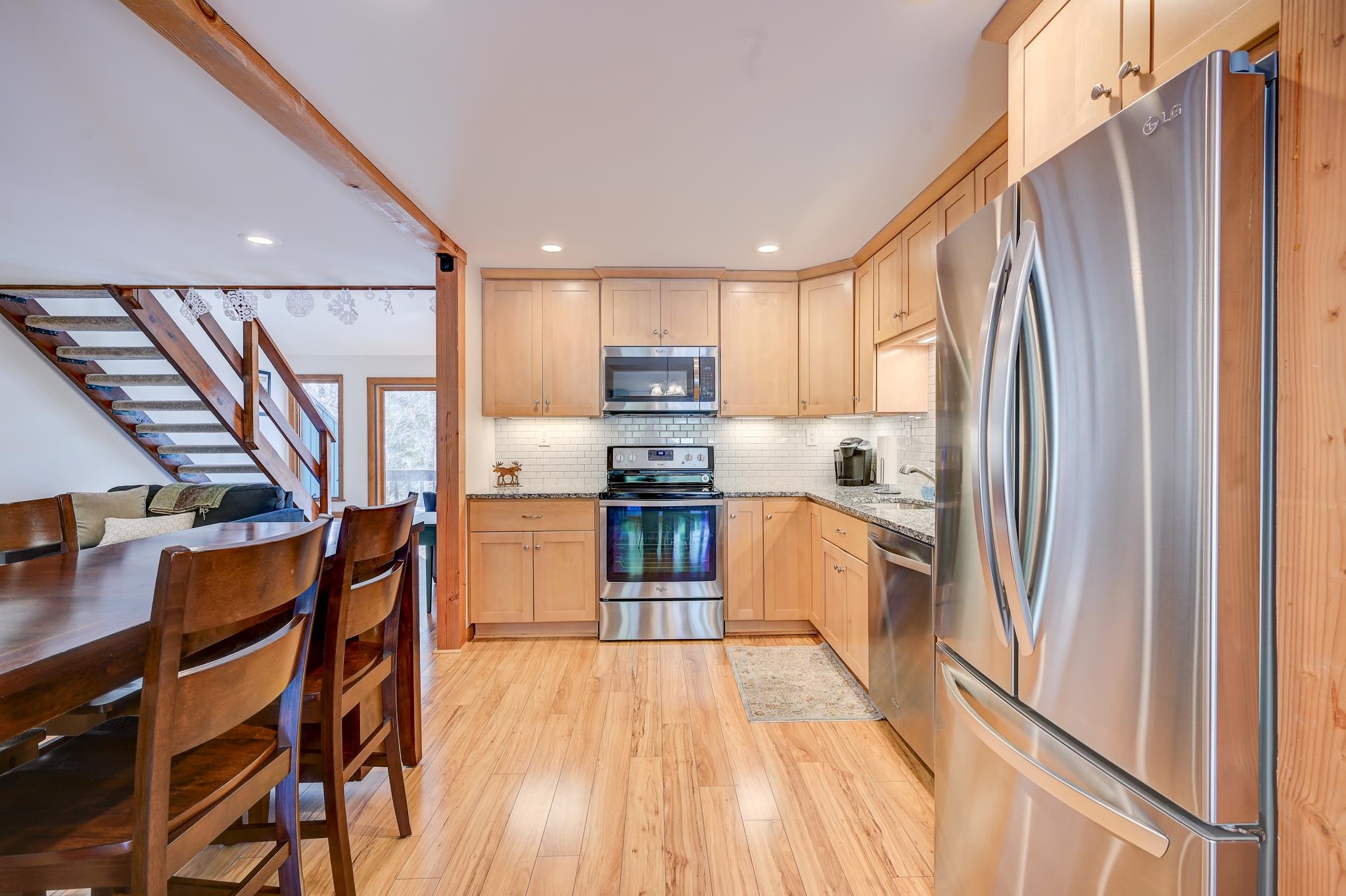Kitchen with light wood cabinets and stainless steel appliances, opening to a dining area and staircase.