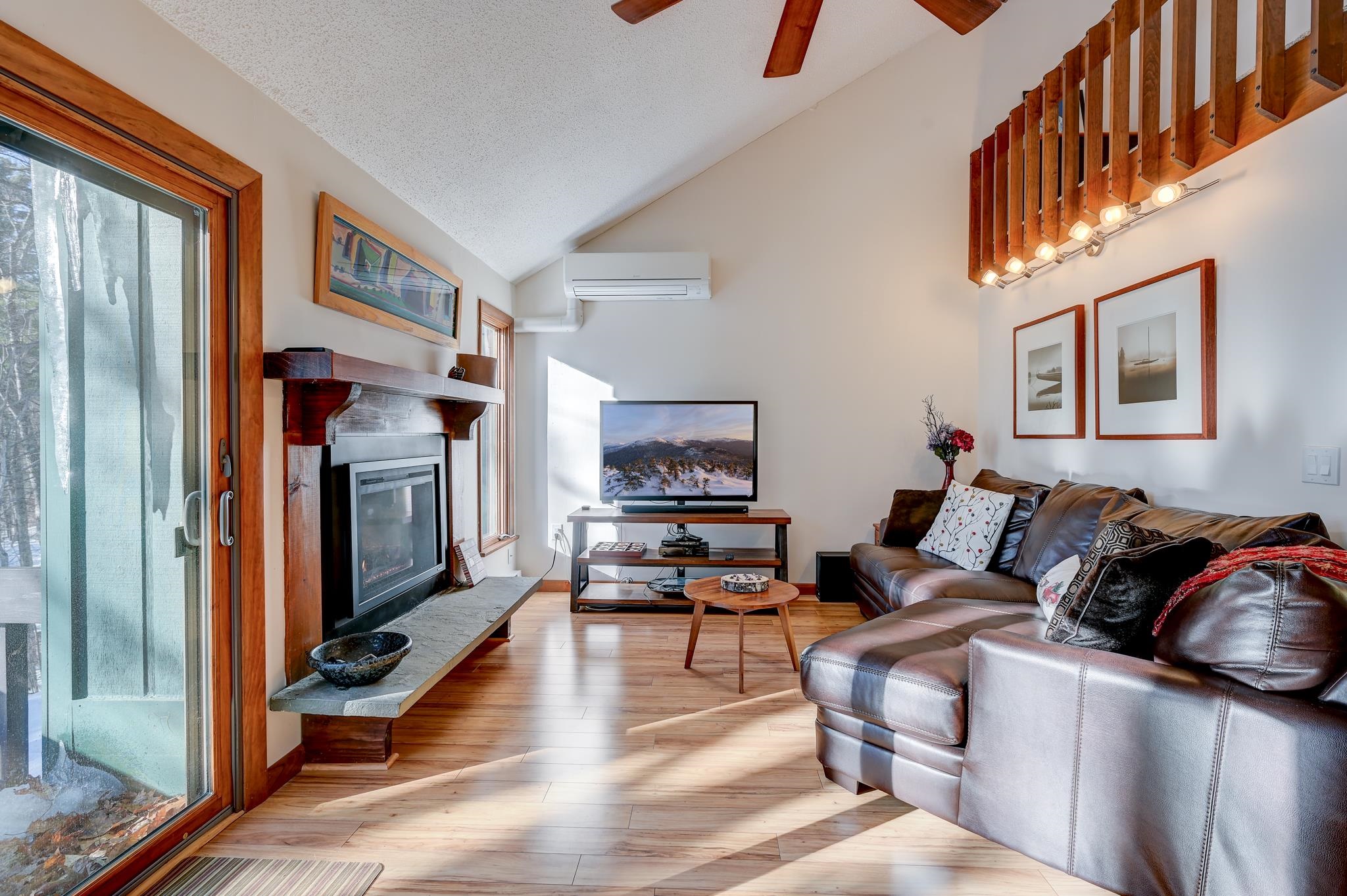 Living room with a stone-surround fireplace, leather sectional, vaulted ceiling, and sliding doors facing snowy trees.