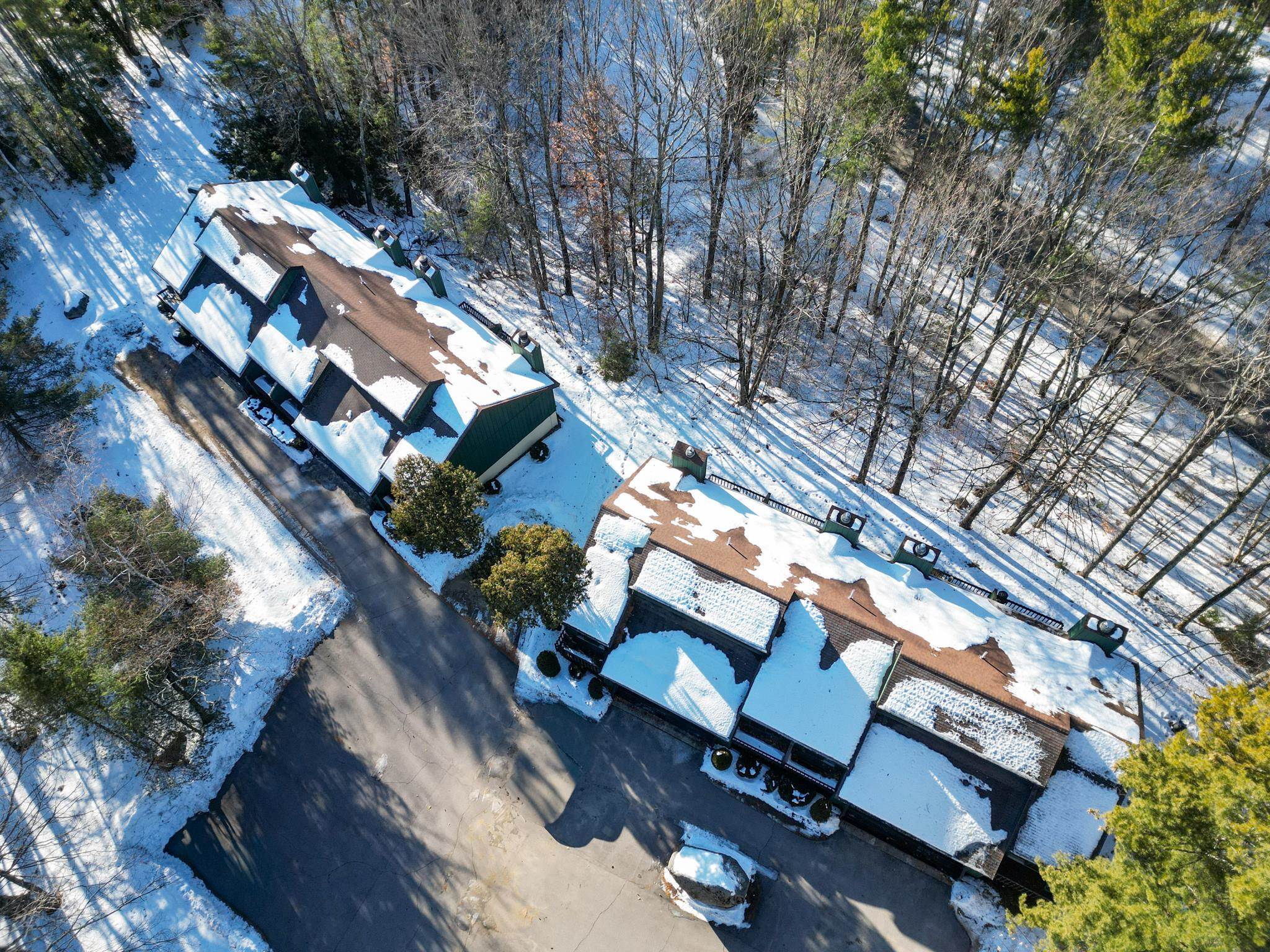 Aerial view of snow-covered condominium buildings beside a paved drive and leafless woods.