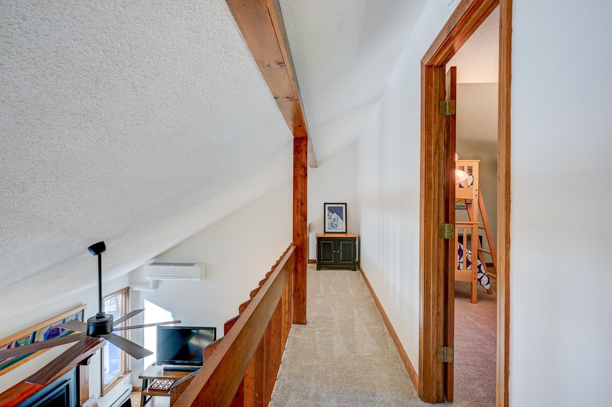 Upstairs hallway with exposed wood beams and railing overlooking the living room, beside an open bunk room.