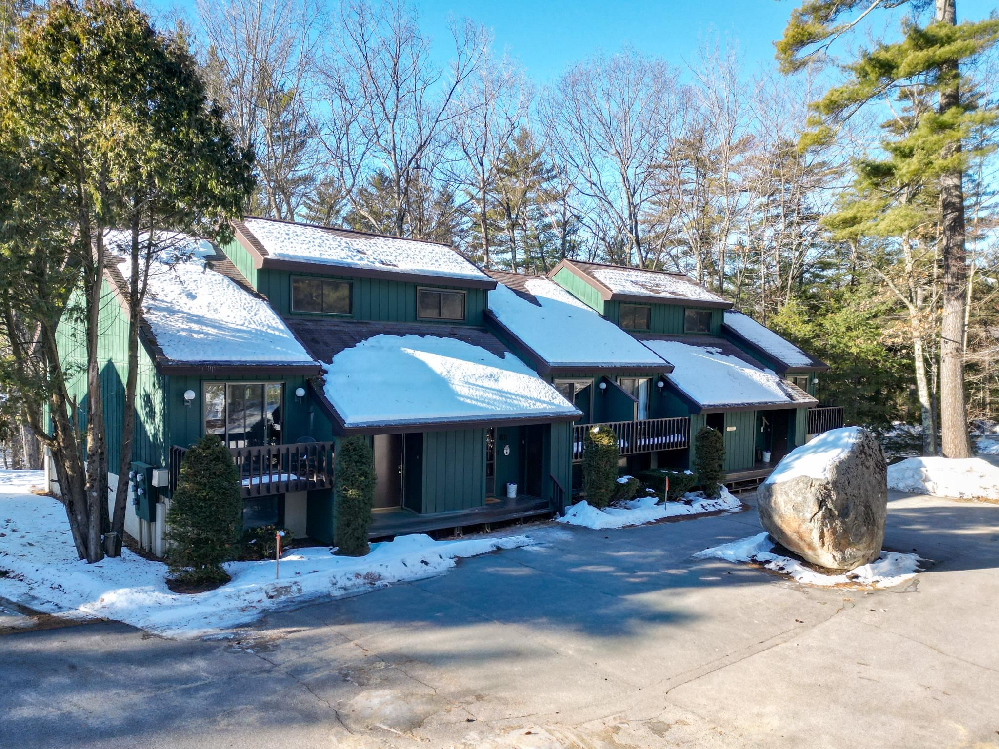Green condominium building with snow-covered roofs, front decks, and a large boulder beside the driveway.