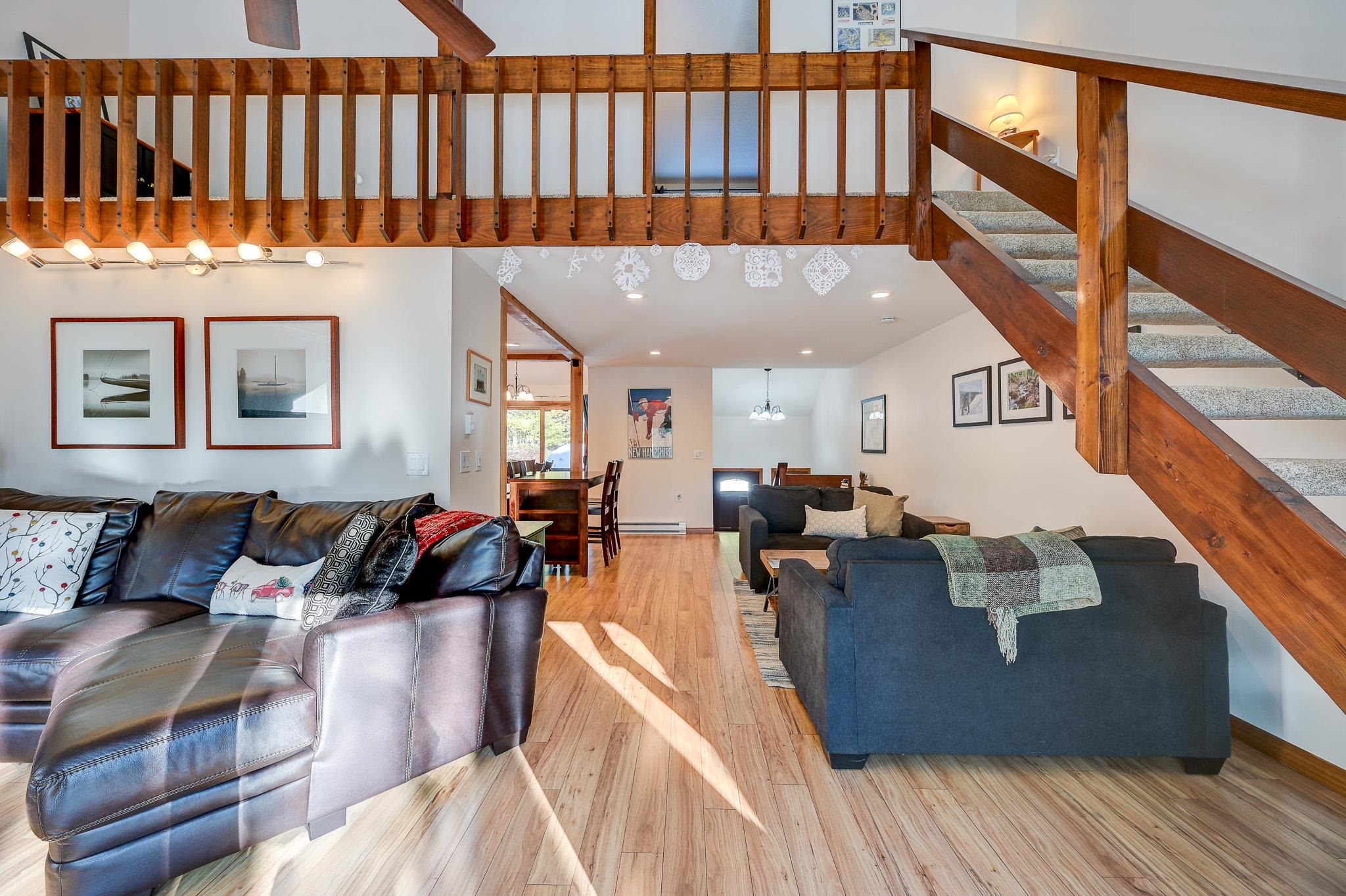 Furnished living room with a wooden loft railing, staircase, sofas, and sunlit wood floors.