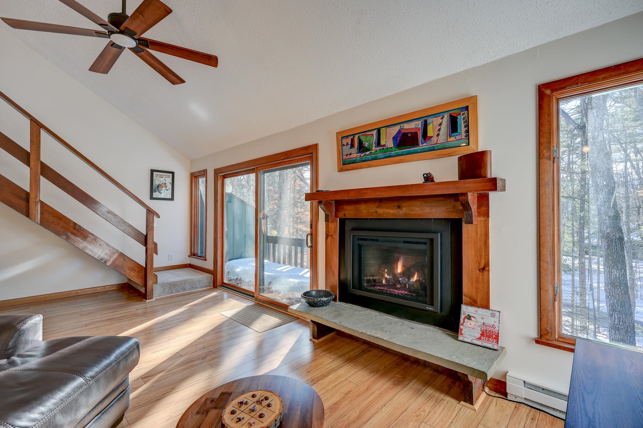 Living room with a large wooden fireplace mantel, hardwood floors, and windows overlooking snowy woods.
