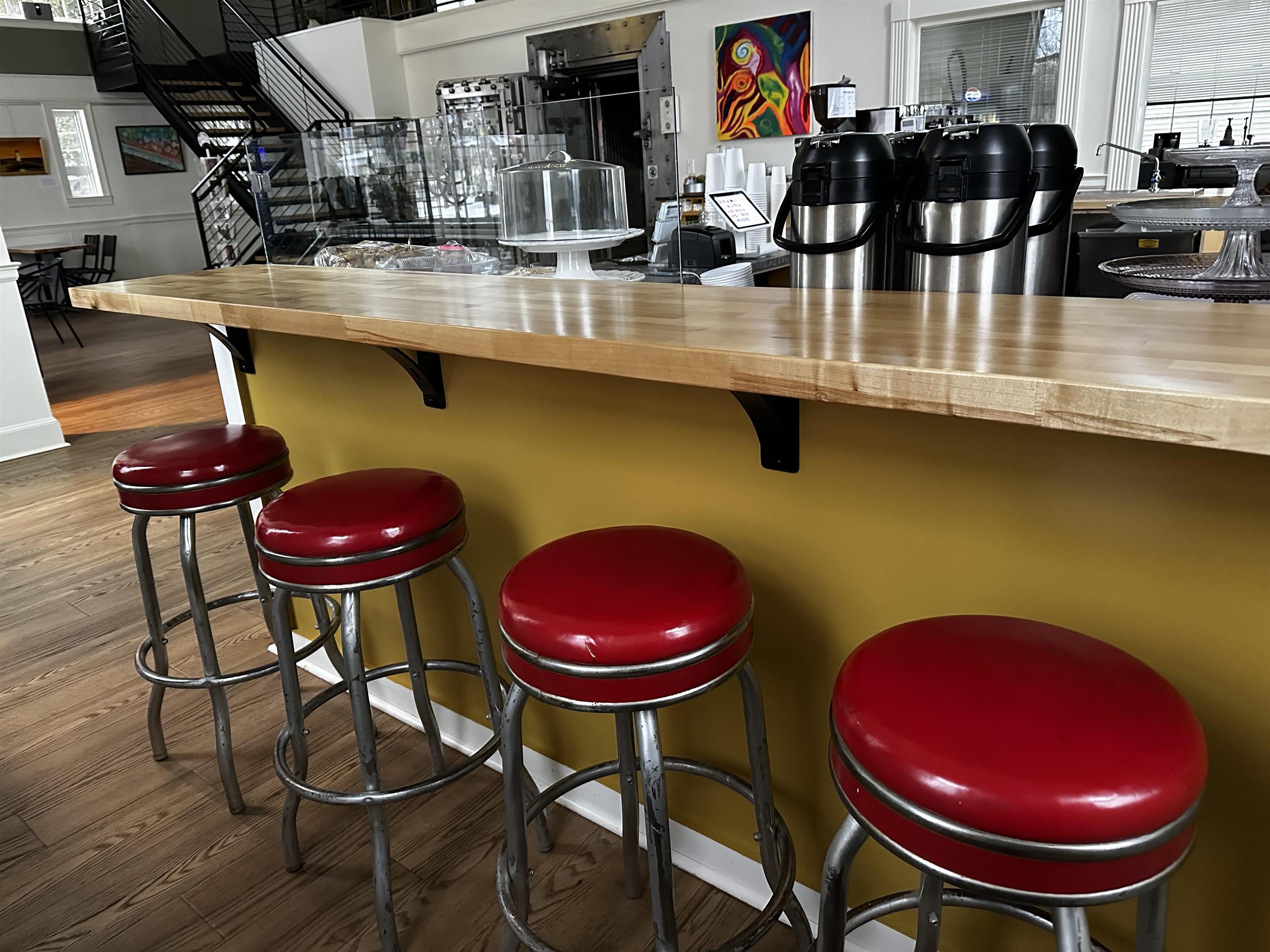 Café counter with four red stools, coffee urns, and a black metal staircase.