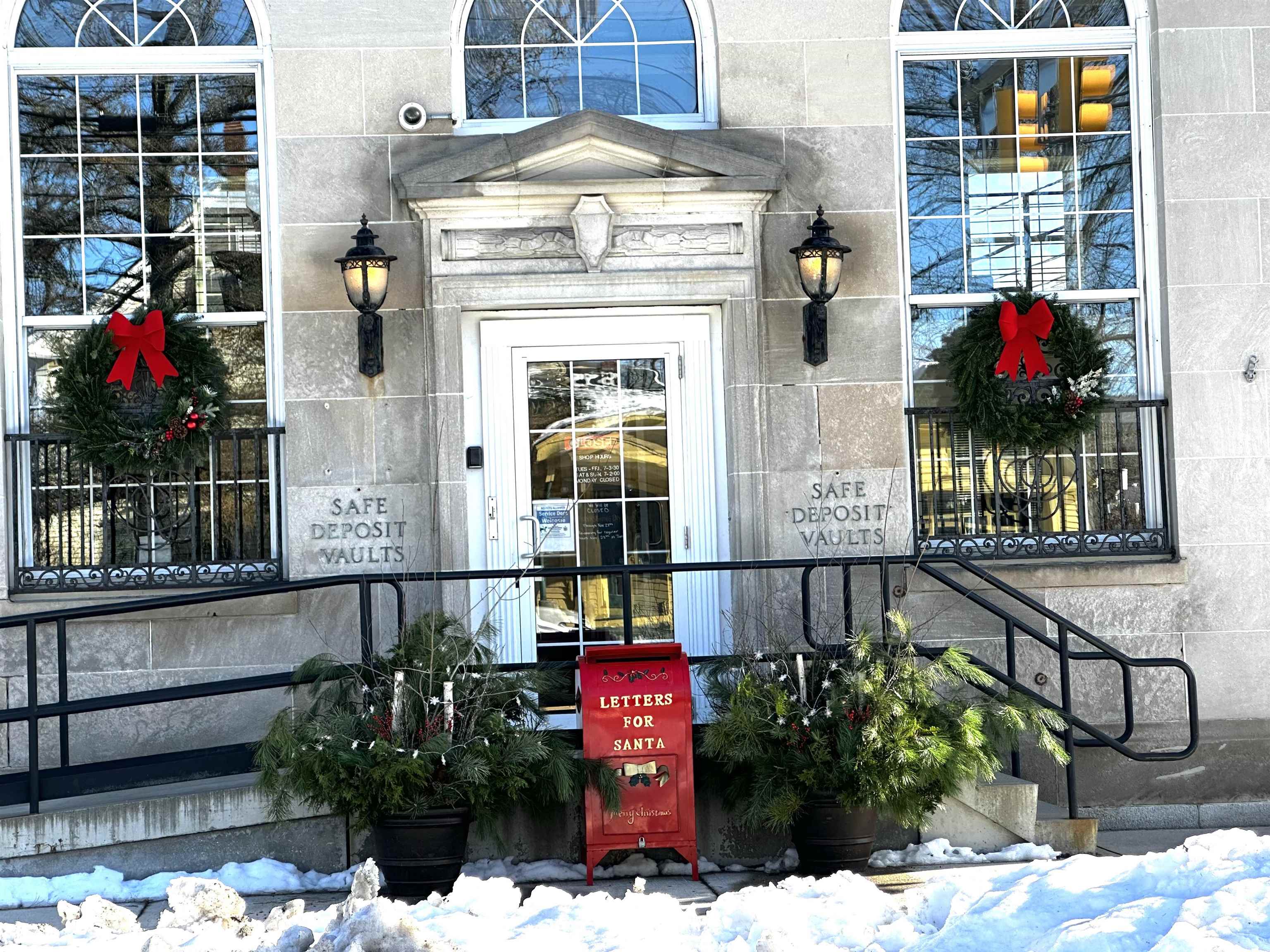 Stone building entrance with arched windows, holiday wreaths, and a red Letters for Santa mailbox in the snow.