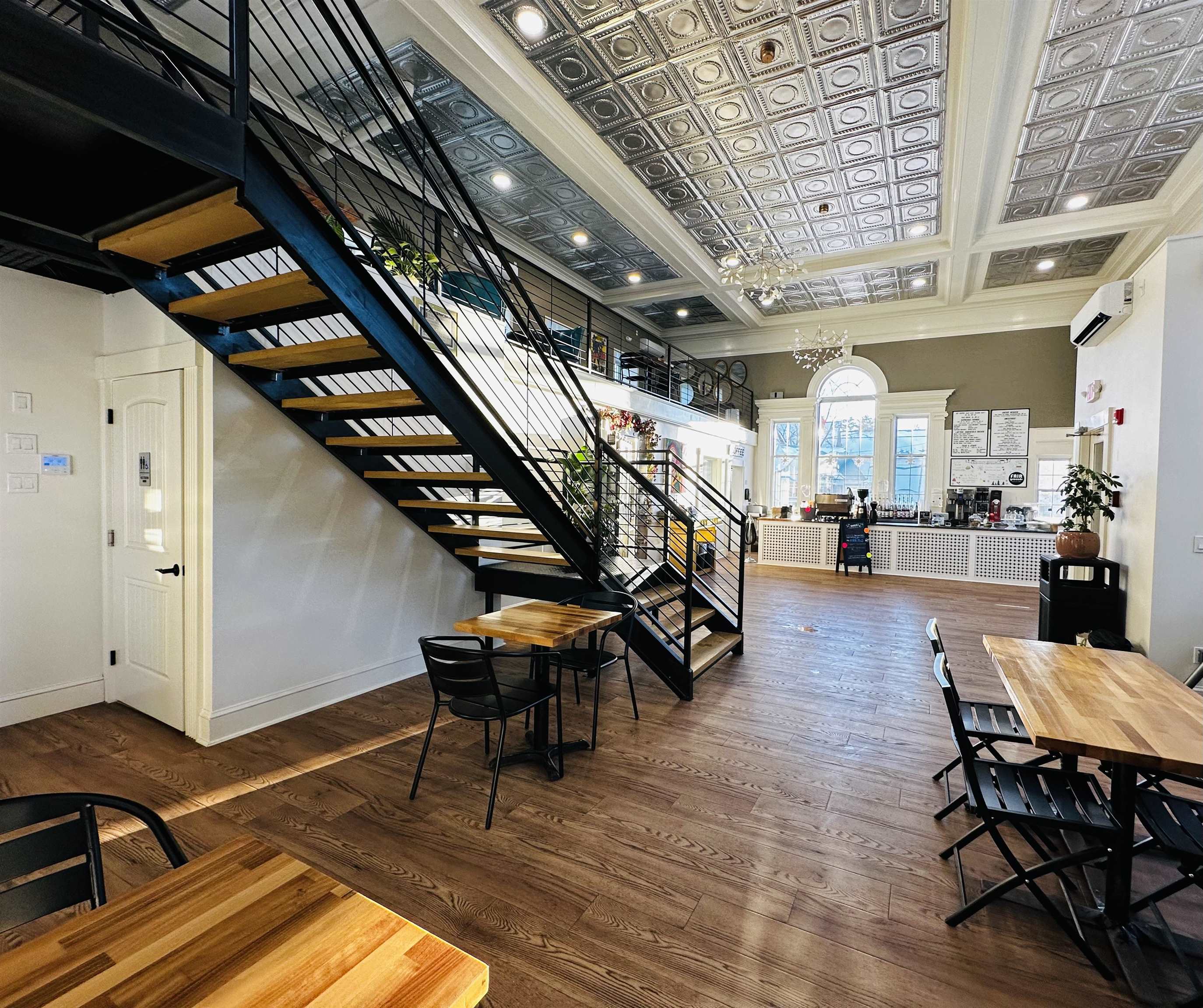 Café interior with a black metal staircase, tin ceiling, wood floors, and a counter beneath tall windows.