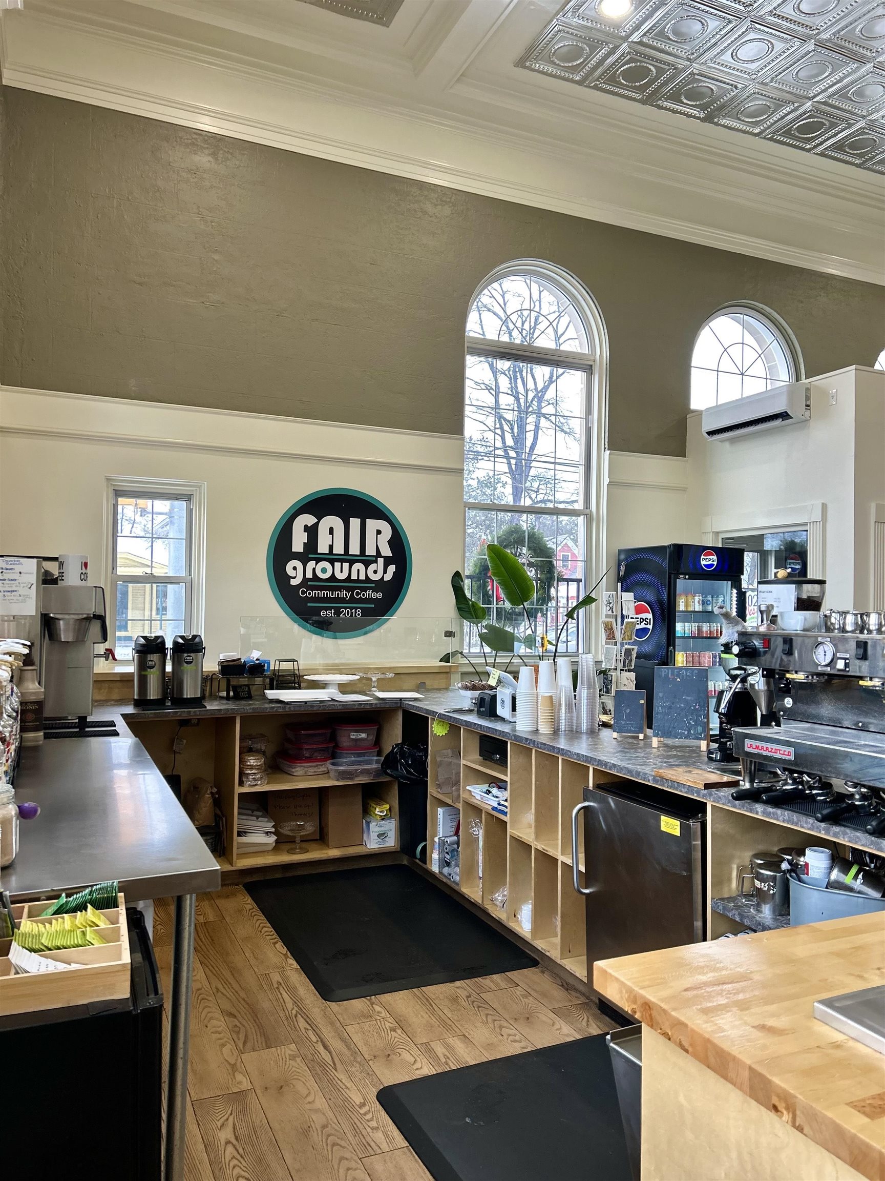 Coffee shop counter beneath tall arched windows, with an ornate ceiling and FAIR grounds sign.