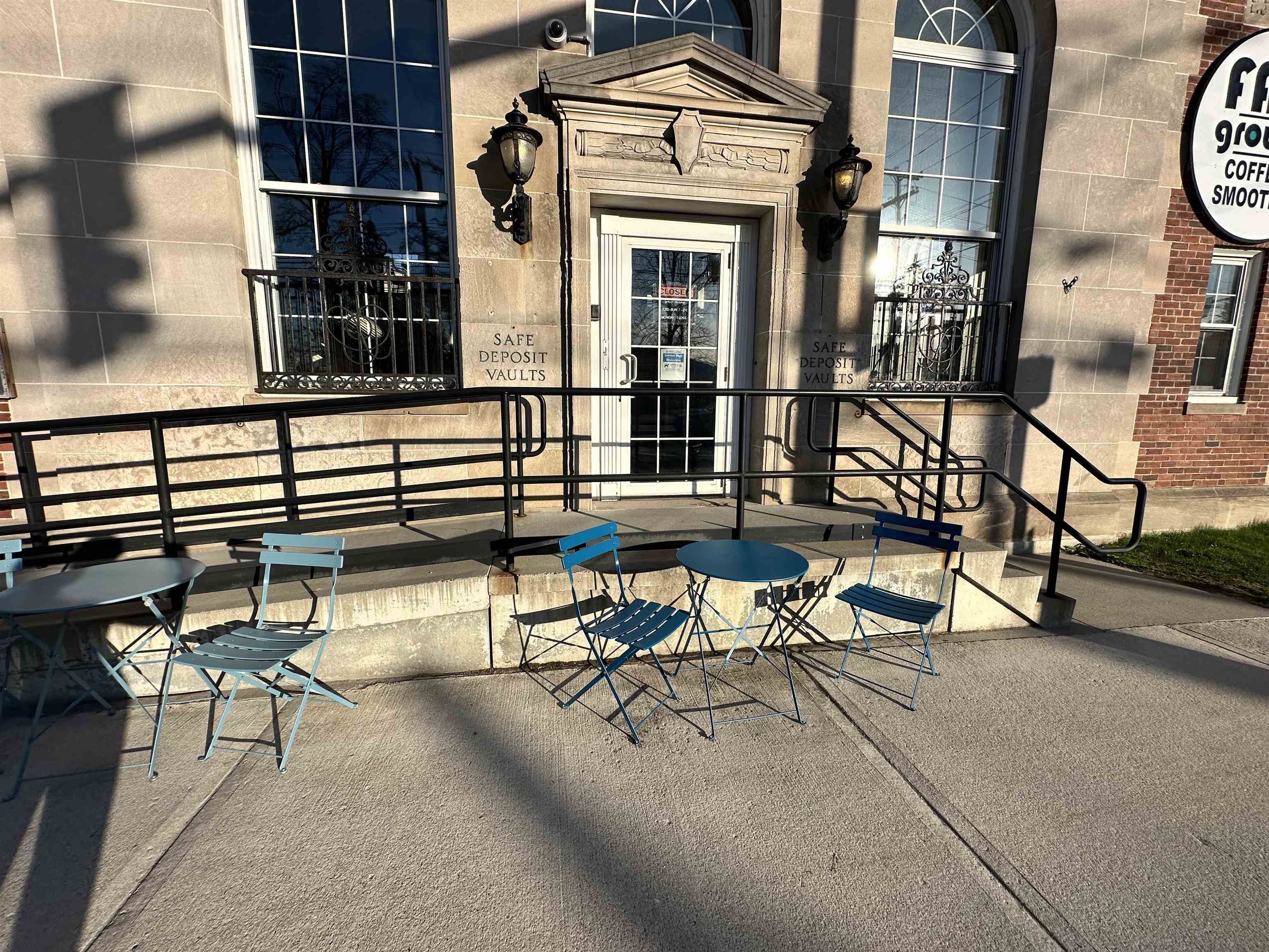 Stone-fronted commercial building entrance with a ramp and blue café seating on the sidewalk.