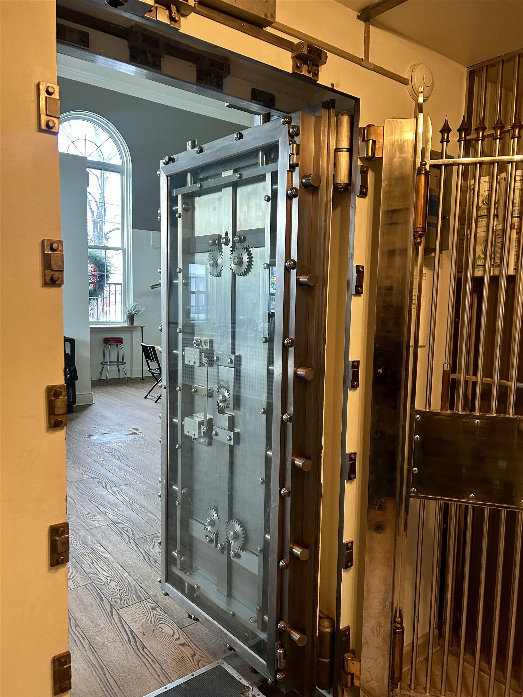 Open glass vault door with exposed gears, revealing a bright room with an arched window and wood-look floors.