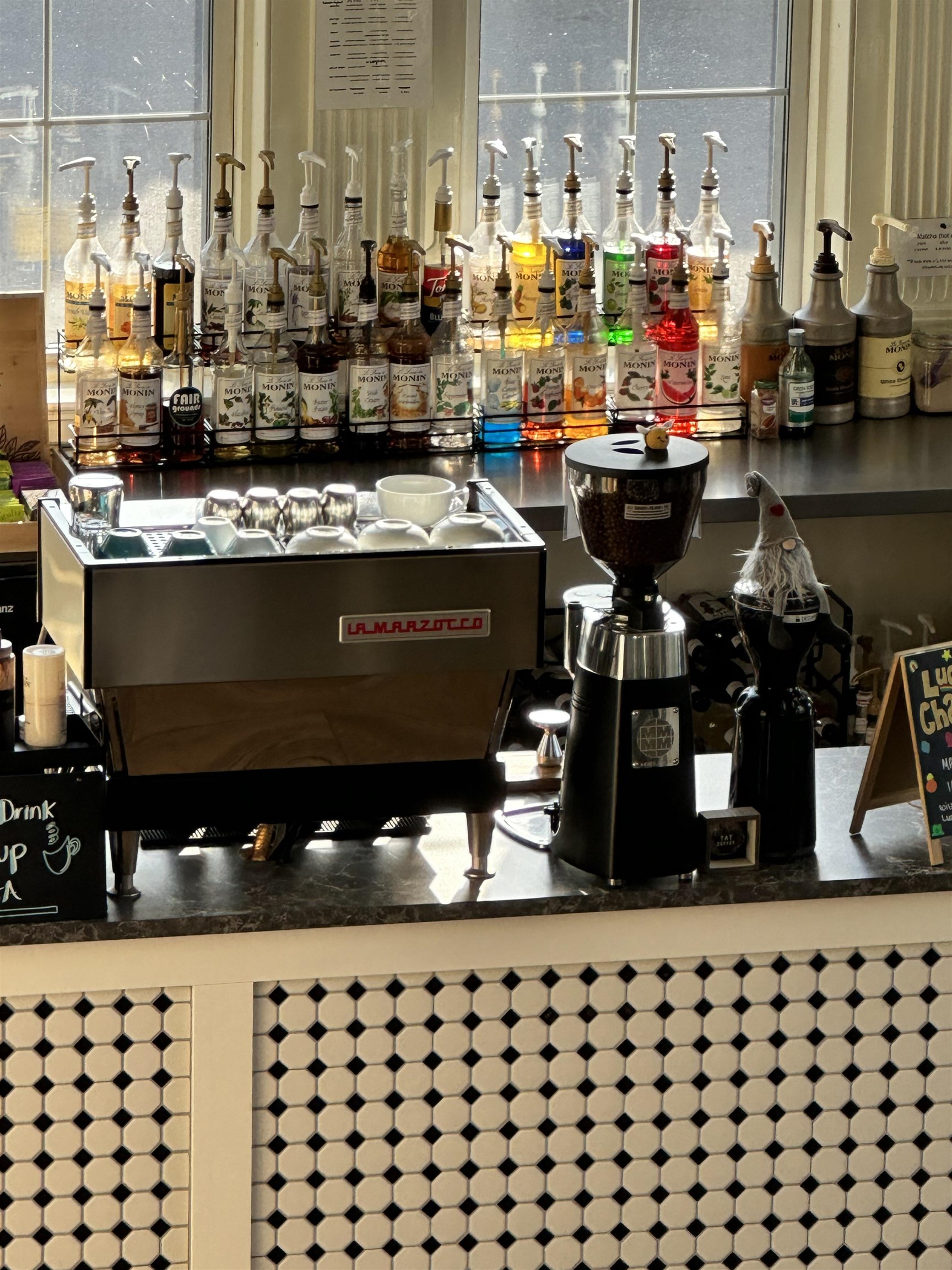 Cafe counter with espresso machine, coffee grinder, and colorful syrup bottles above geometric tilework.