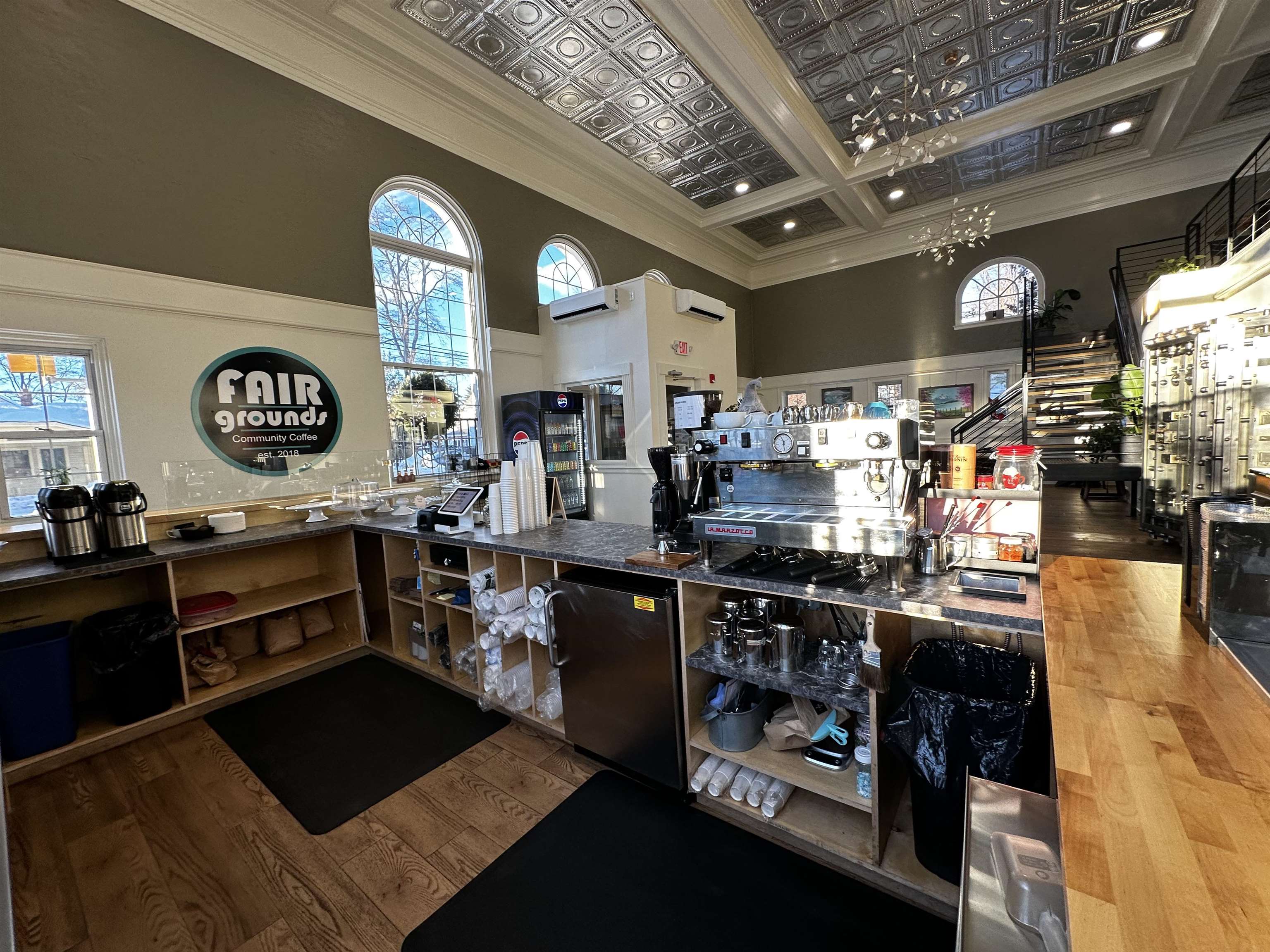Coffee shop counter with espresso equipment beneath a decorative tin ceiling and arched windows.