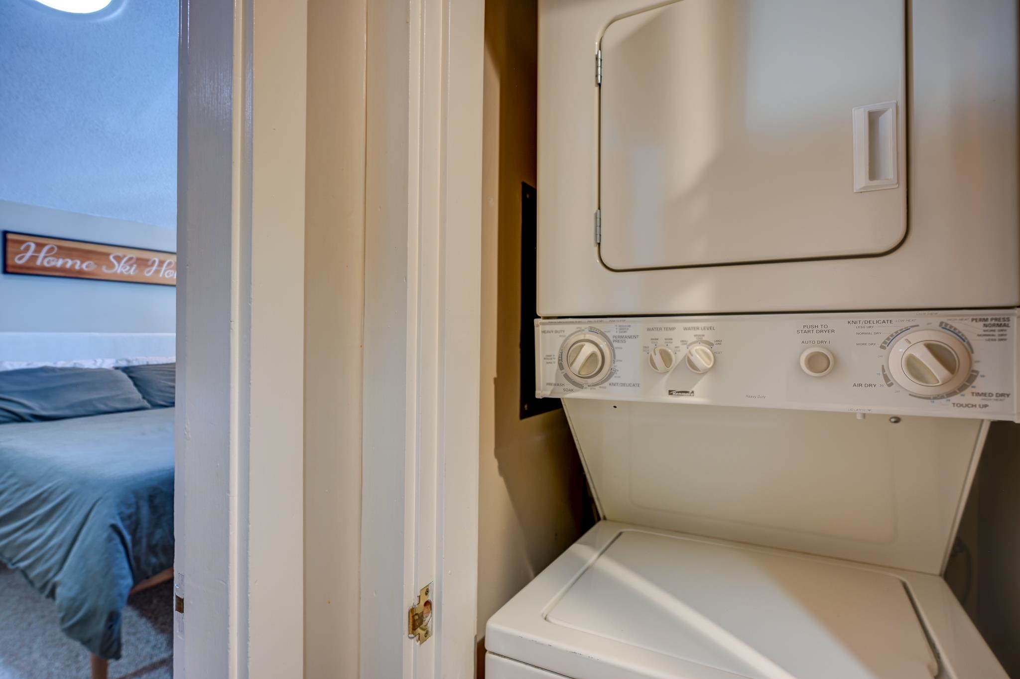 Stacked washer and dryer in a closet beside a bedroom with blue bedding.
