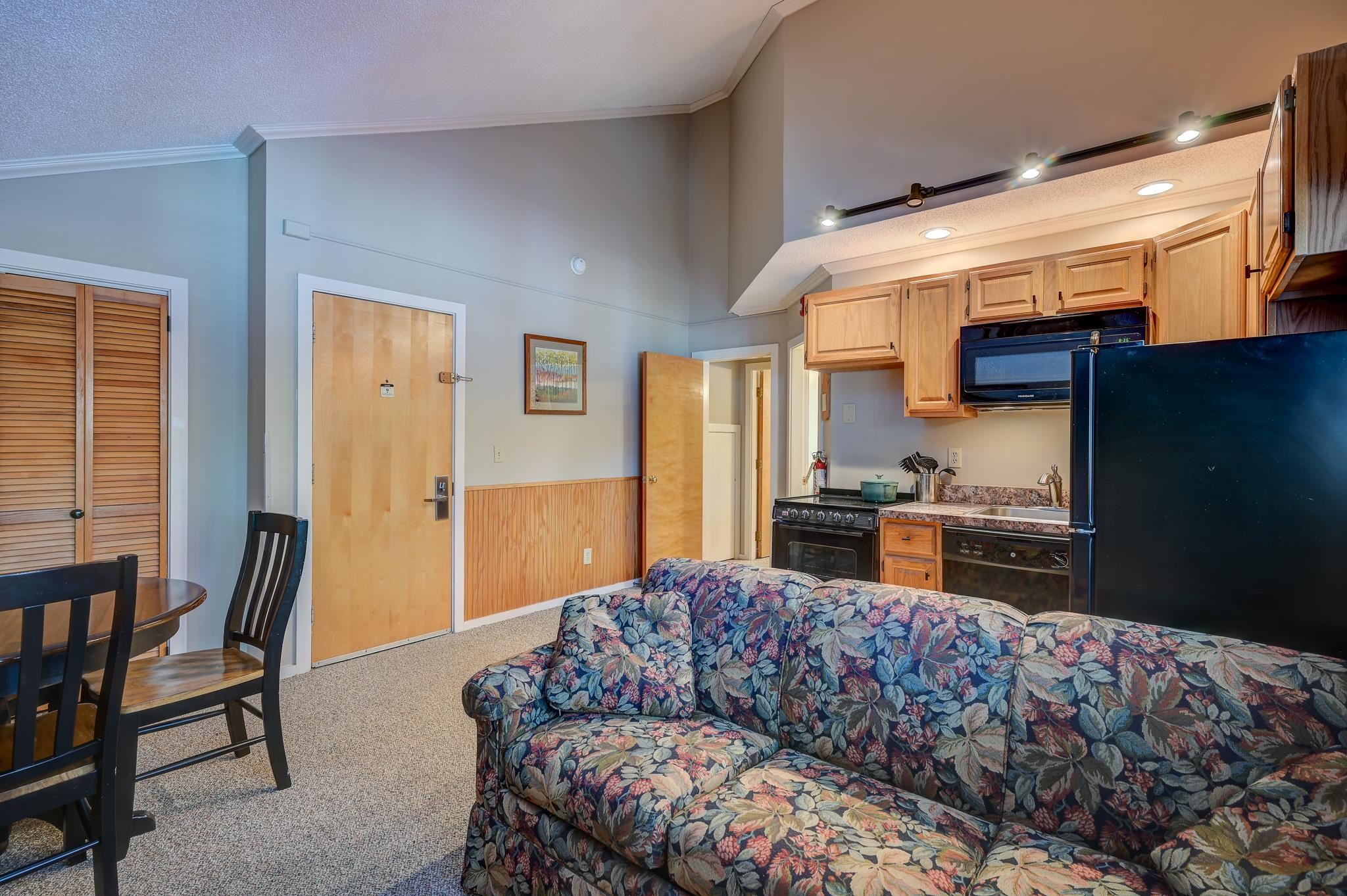 Open-plan living area with a floral sofa, dining table, and compact kitchen with wood cabinets.