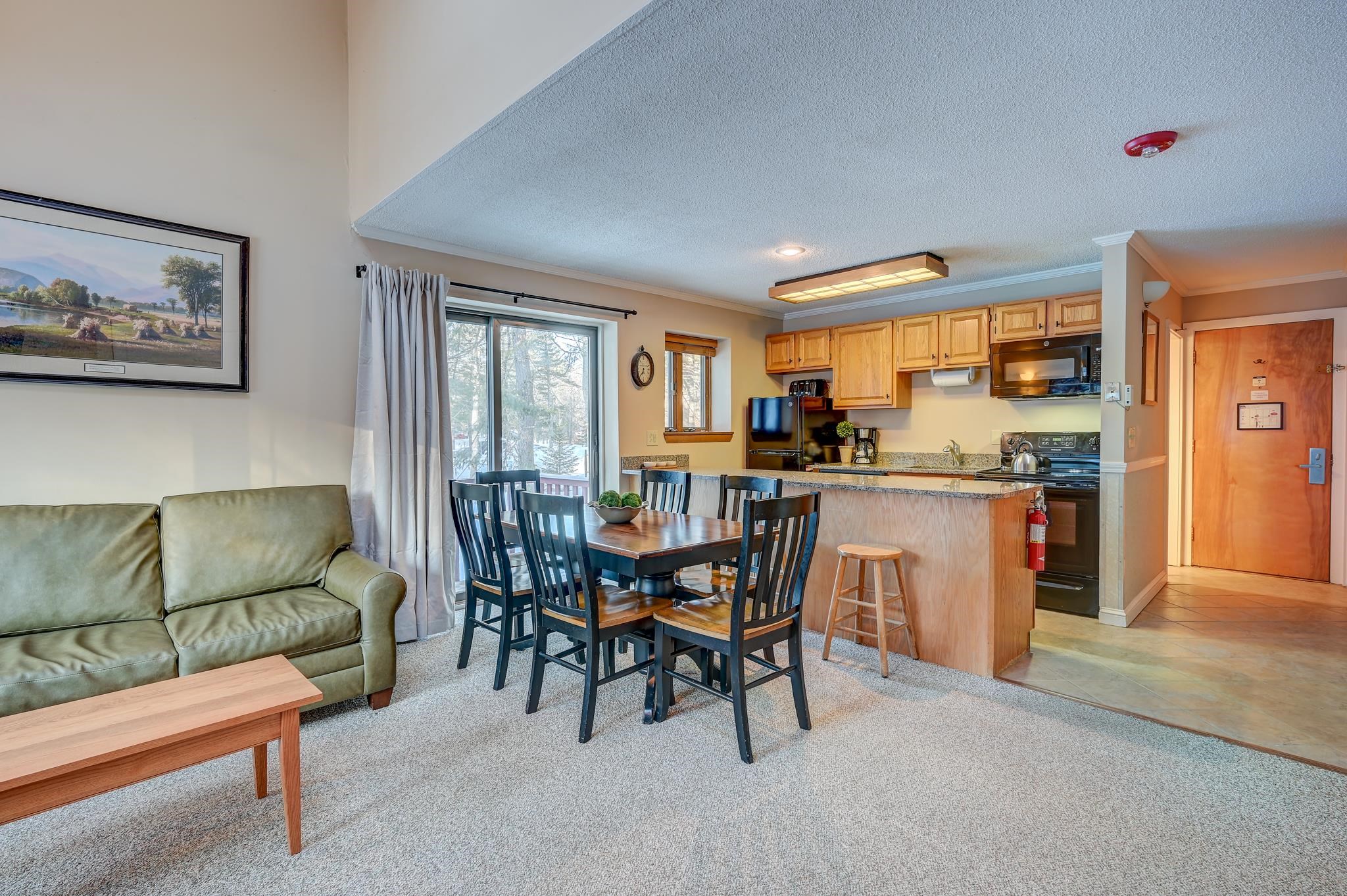 Open-plan condo living and kitchen area with wood cabinets, granite counters, and a dining table by snowy windows.