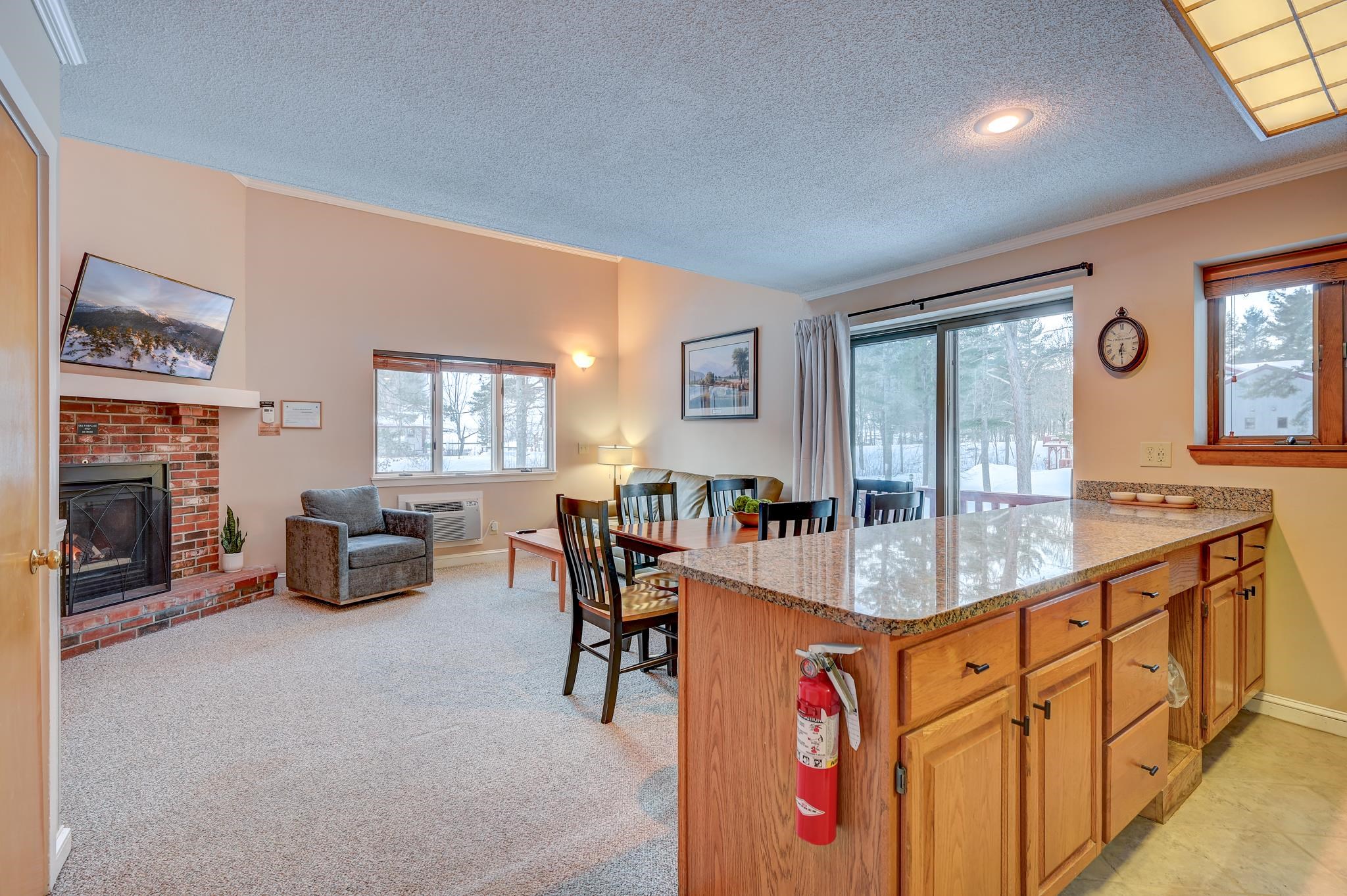 Open-plan condo living and dining area with a granite kitchen peninsula, brick fireplace, and snowy views.