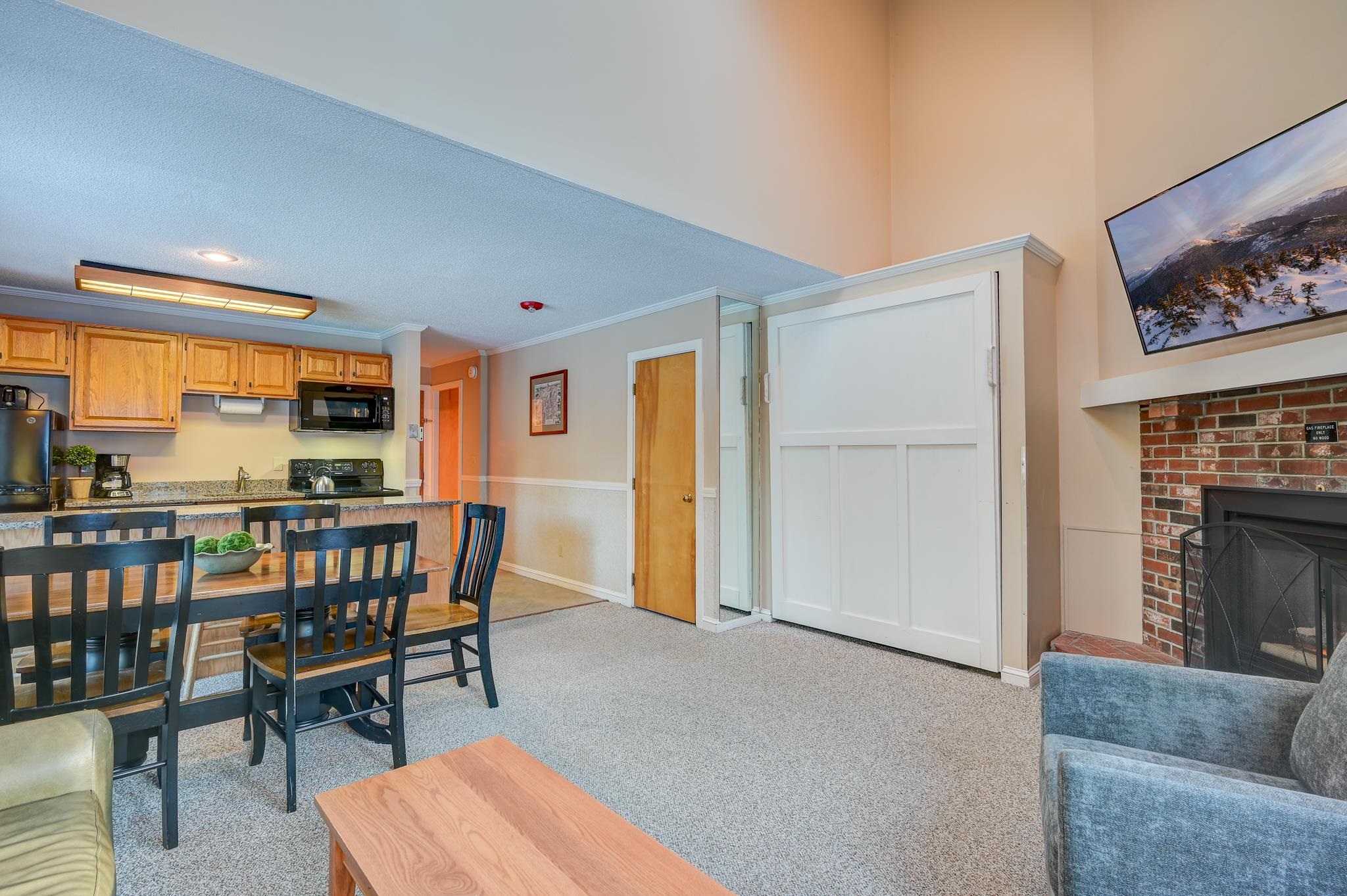 Open-plan living and dining area with a brick fireplace, carpeted floor, and adjoining wood-cabinet kitchen.