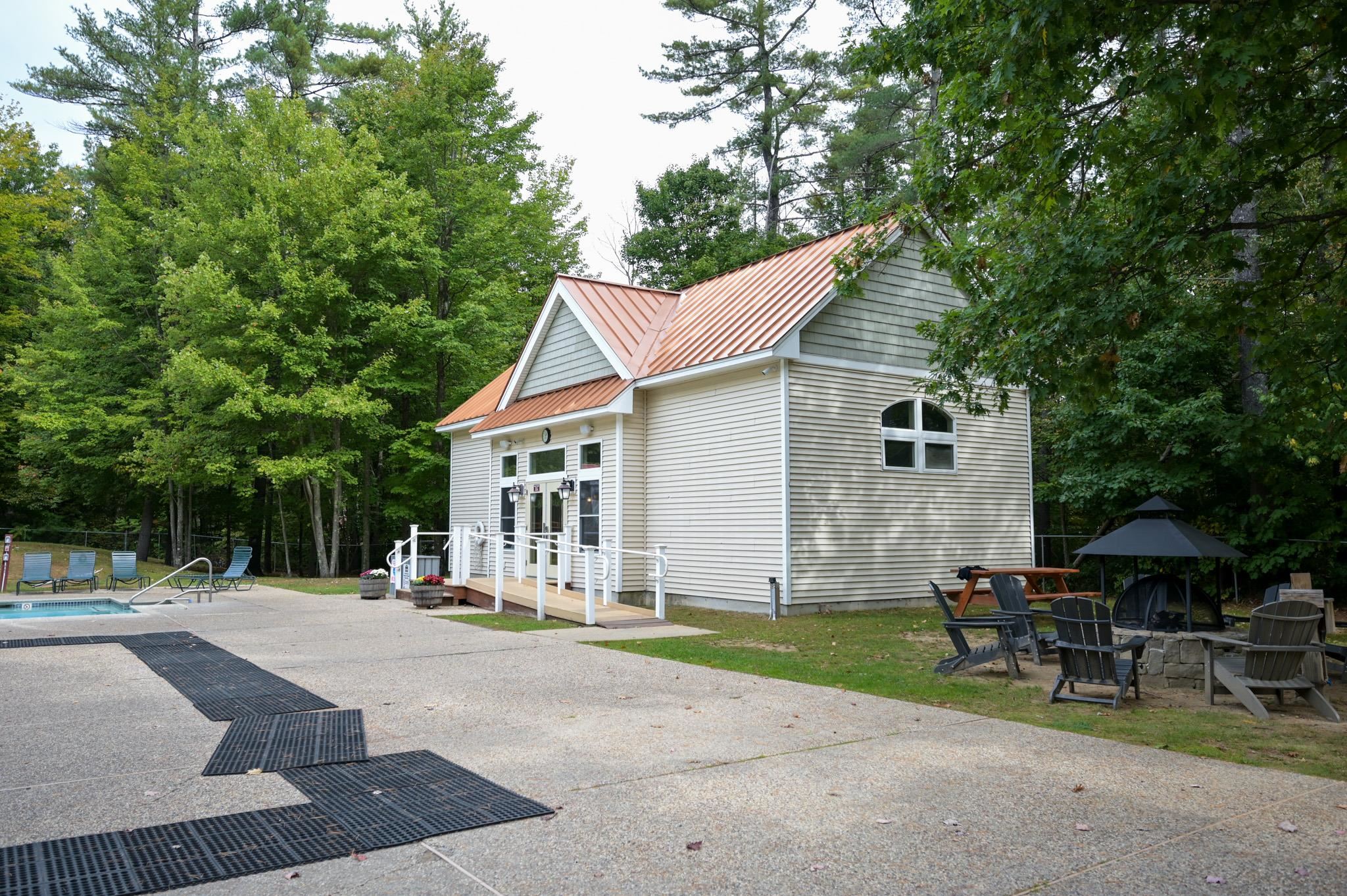 Light-sided building with copper-colored metal roofs beside a pool, ramp, and wooded fire pit area.