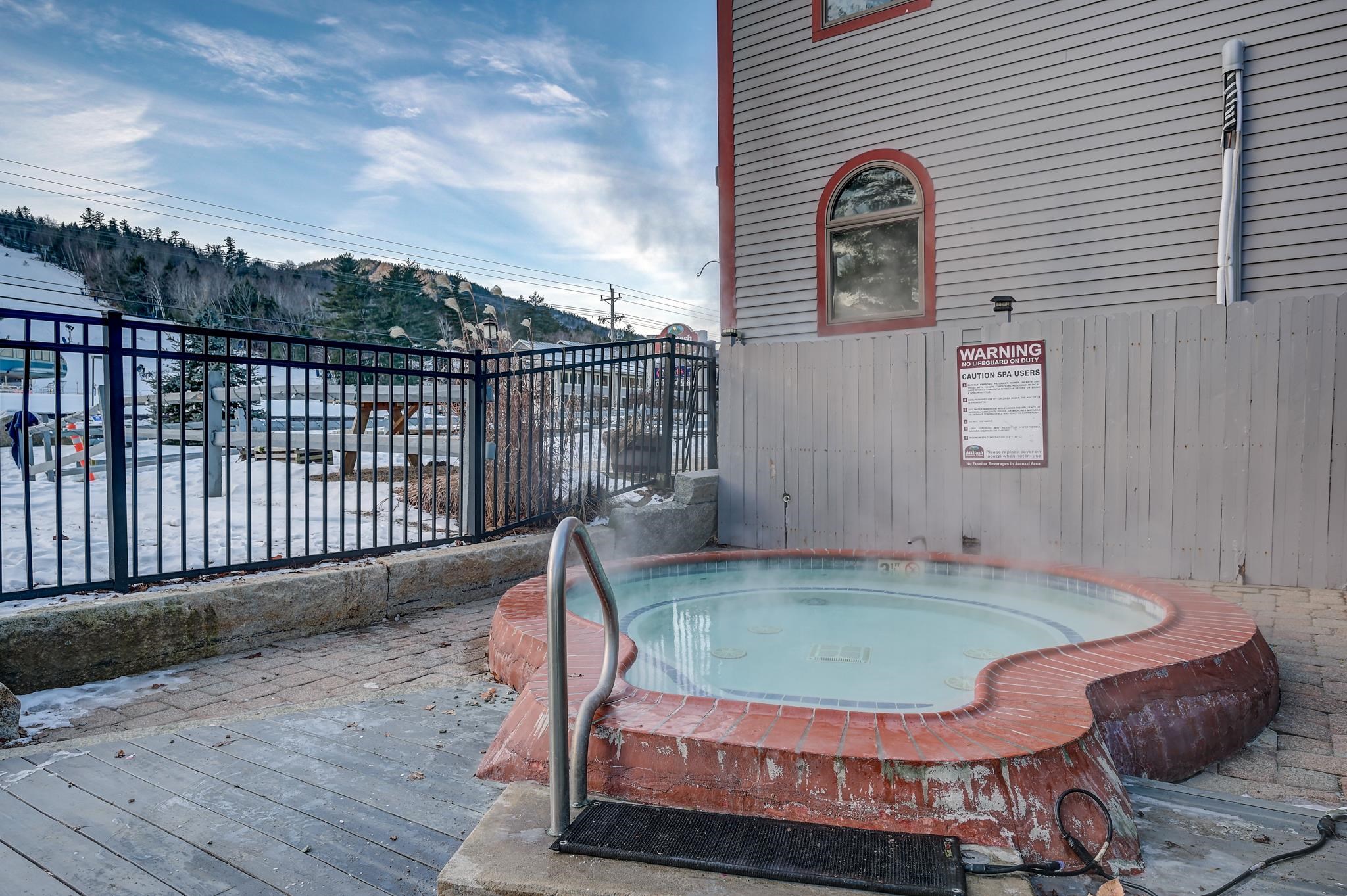 Outdoor hot tub beside a building, with snow and a wooded mountain slope beyond the fence.