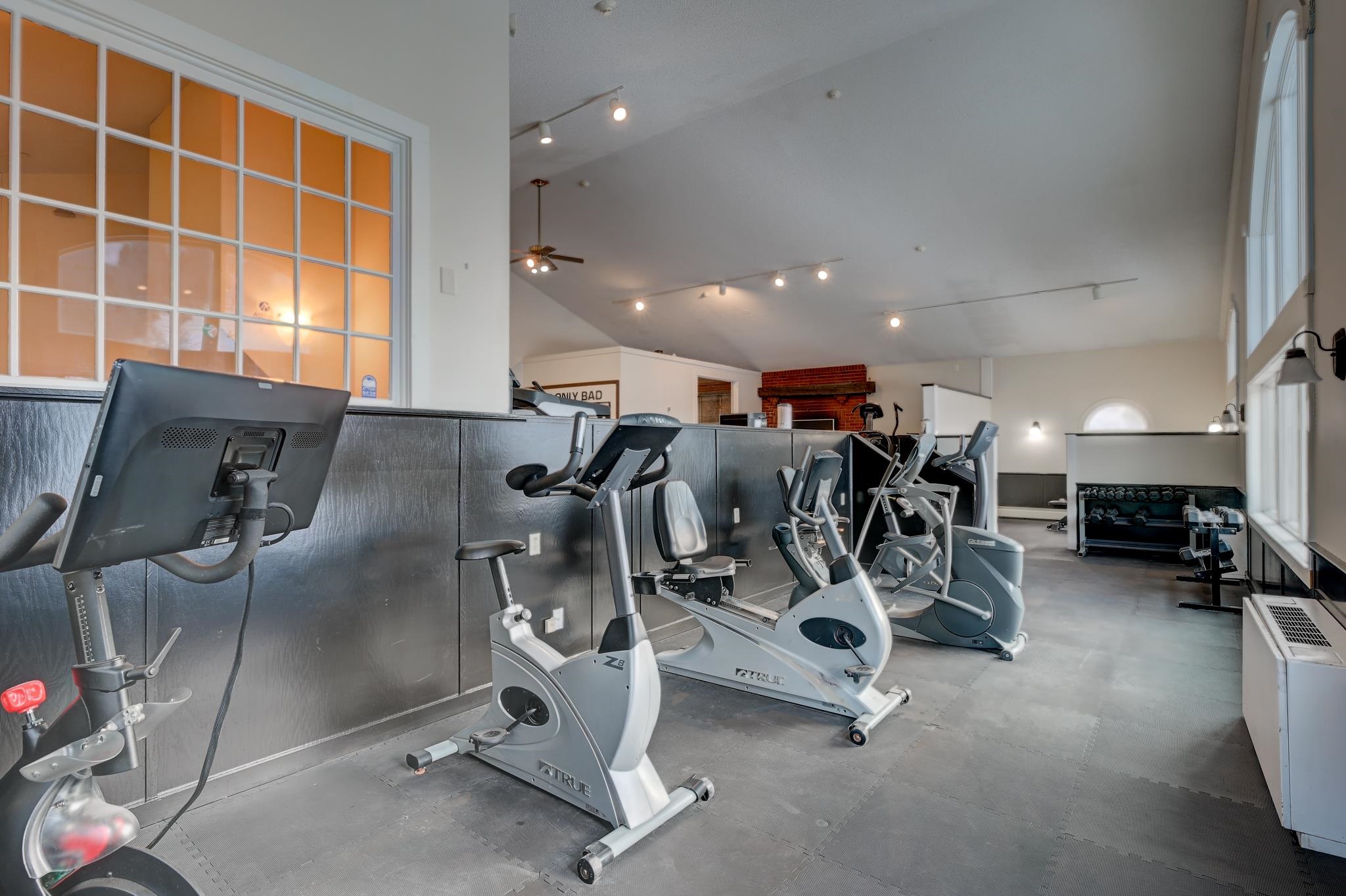 Fitness center with rows of exercise machines beneath a high sloped ceiling and beside large windows.