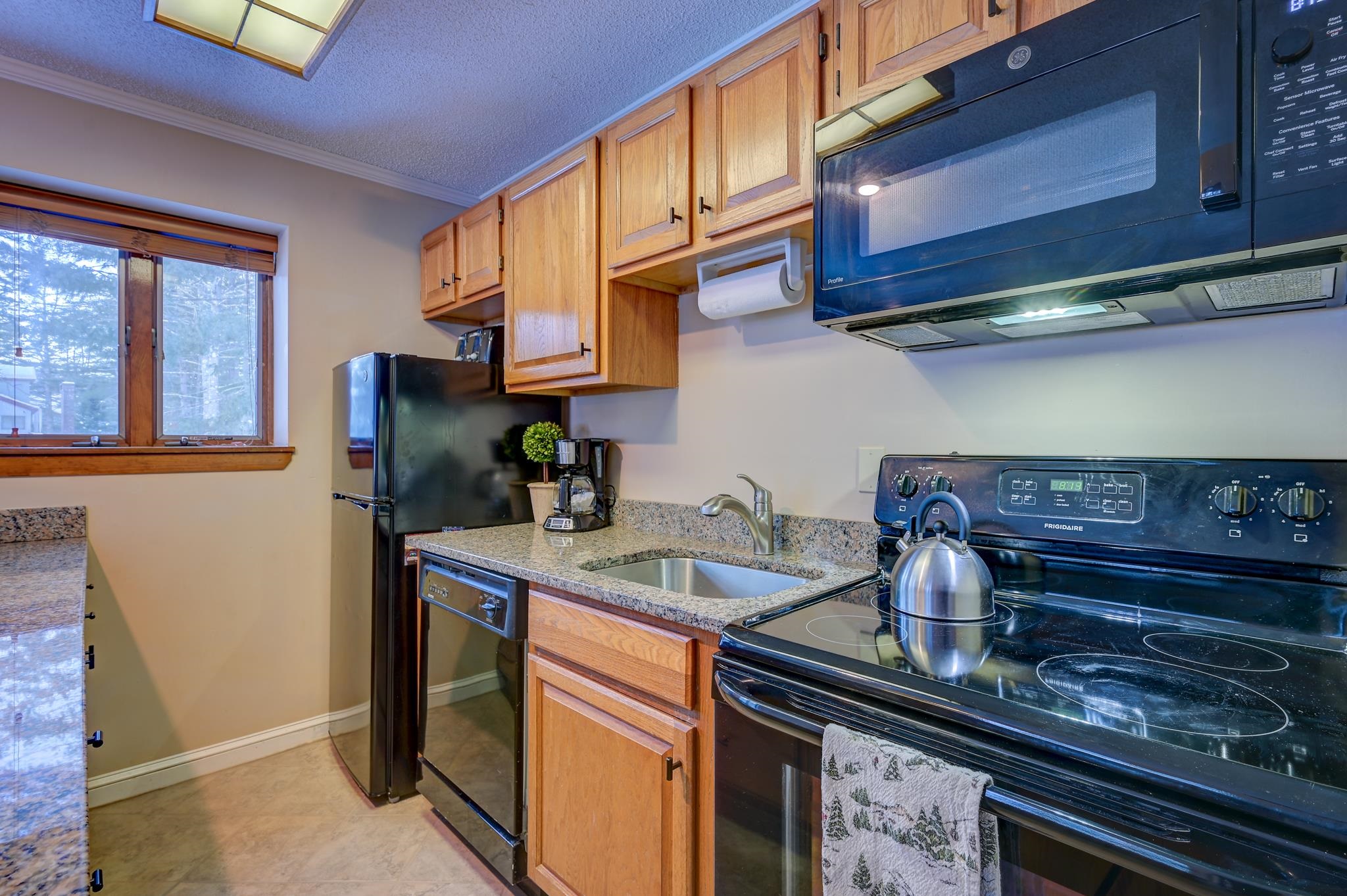 Kitchen with wood cabinets, granite counters, black appliances, and a window overlooking snowy trees.