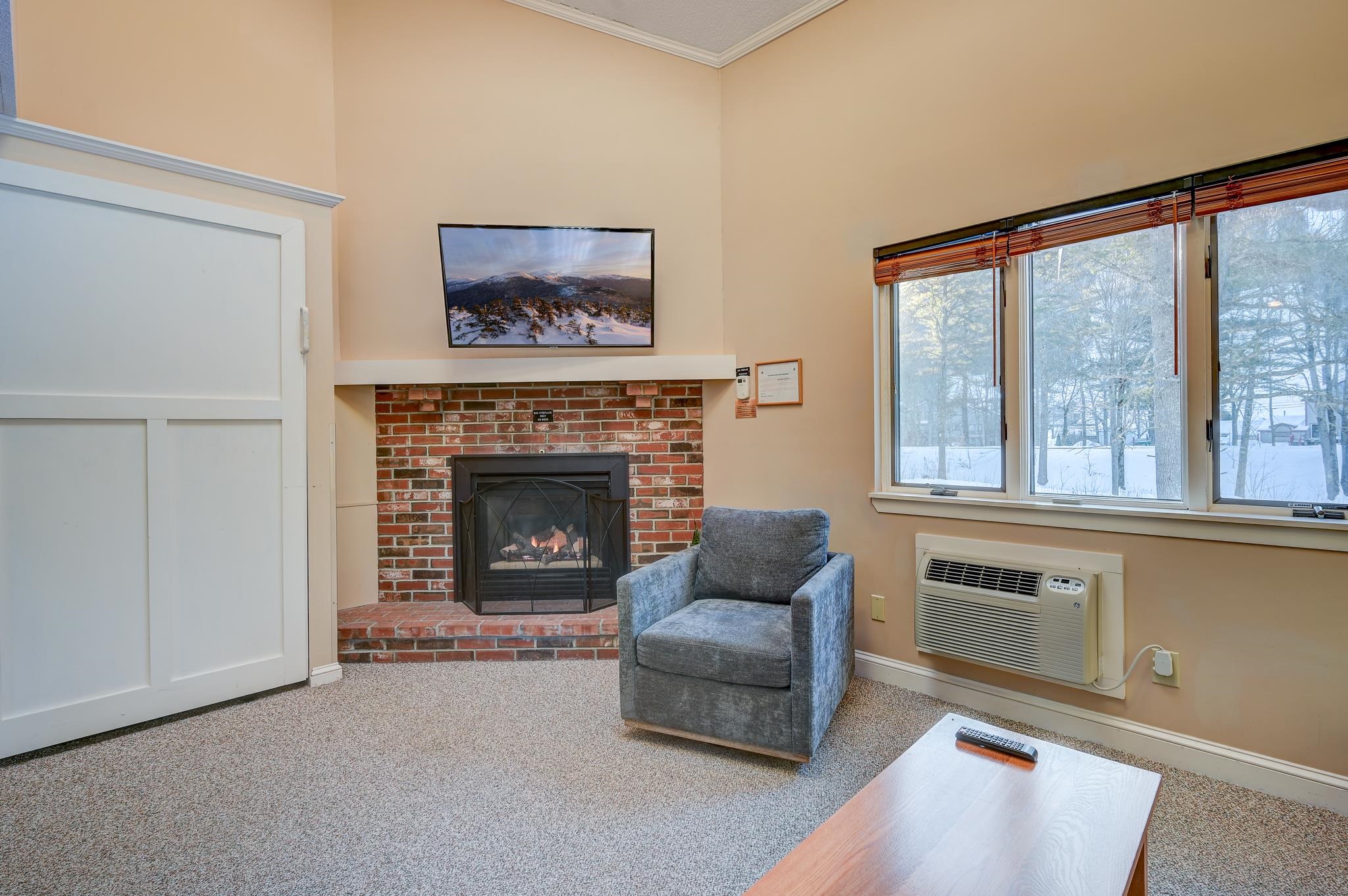 Living room with a brick fireplace, gray armchair, and wide window overlooking a snowy yard.
