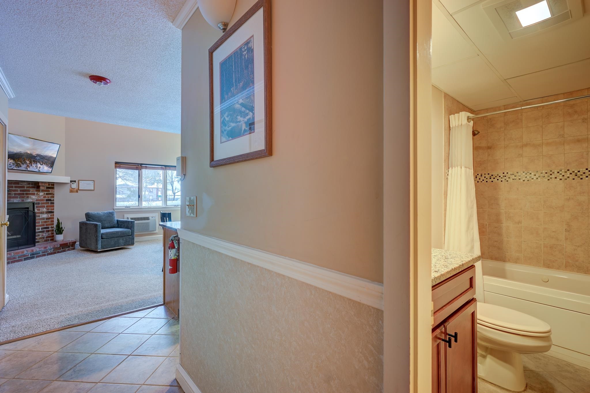 Condo hallway between a tiled bathroom and living room with a brick fireplace and blue armchair.