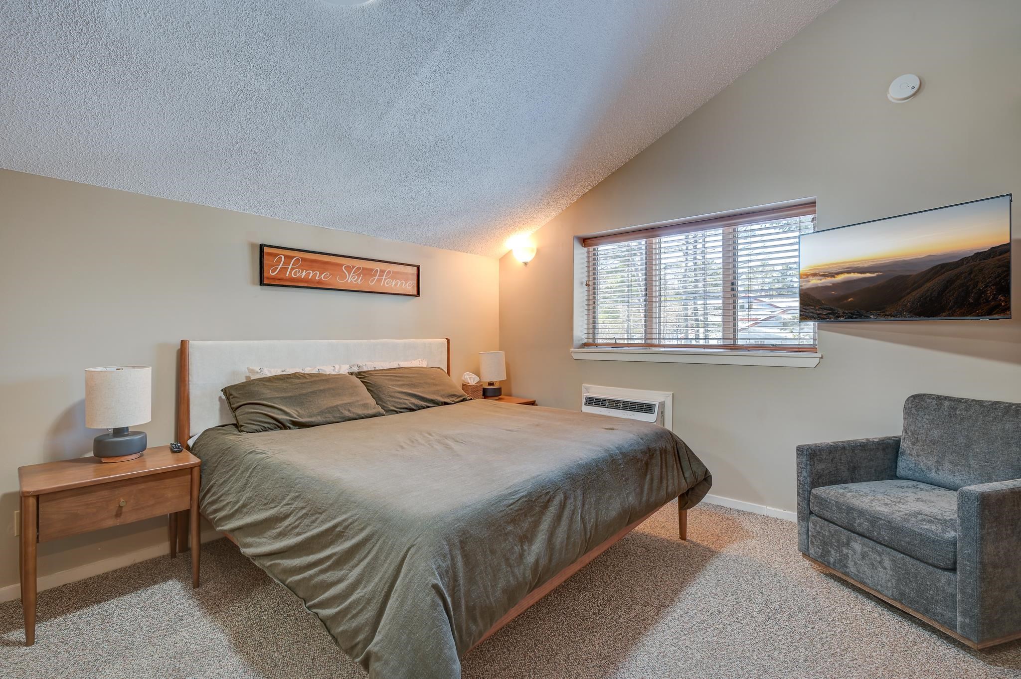 Furnished bedroom with a large bed, gray armchair, and “Home Ski Home” sign above the headboard.