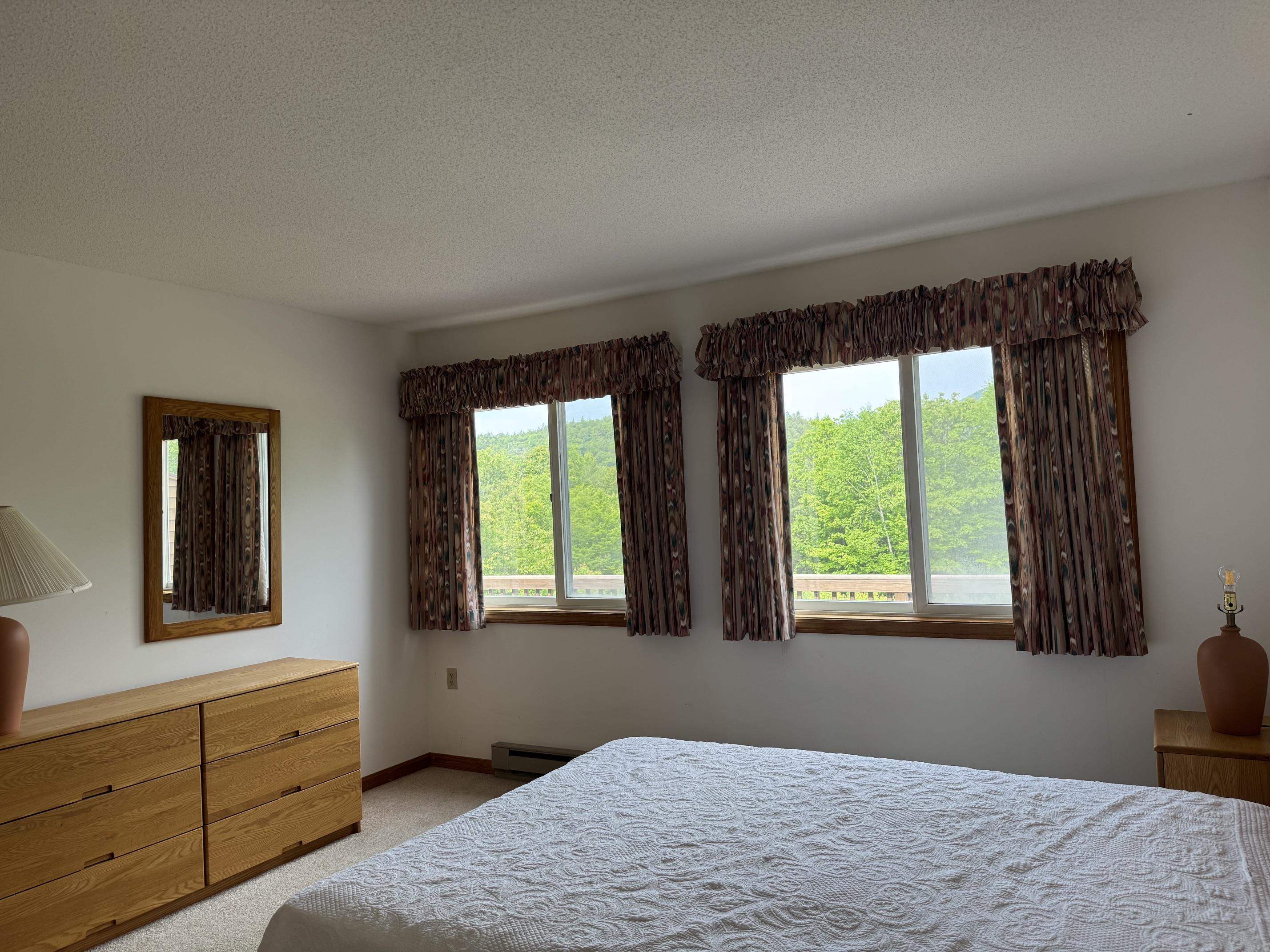 Bedroom with a large bed and windows overlooking leafy trees and a wooded hillside.