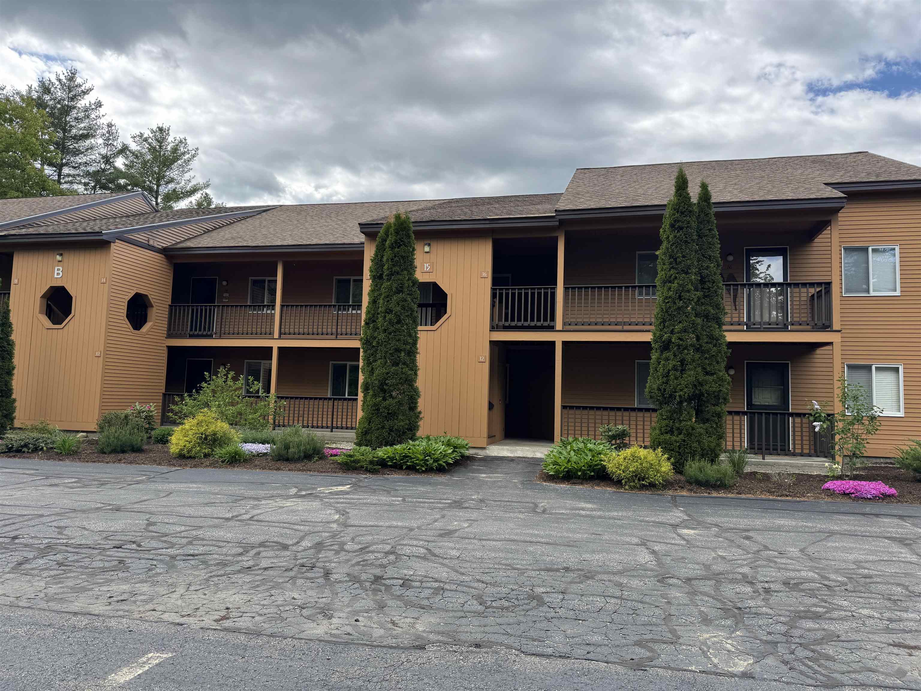 Two-story brown condominium building with covered balconies, evergreen trees, and landscaped flower beds.