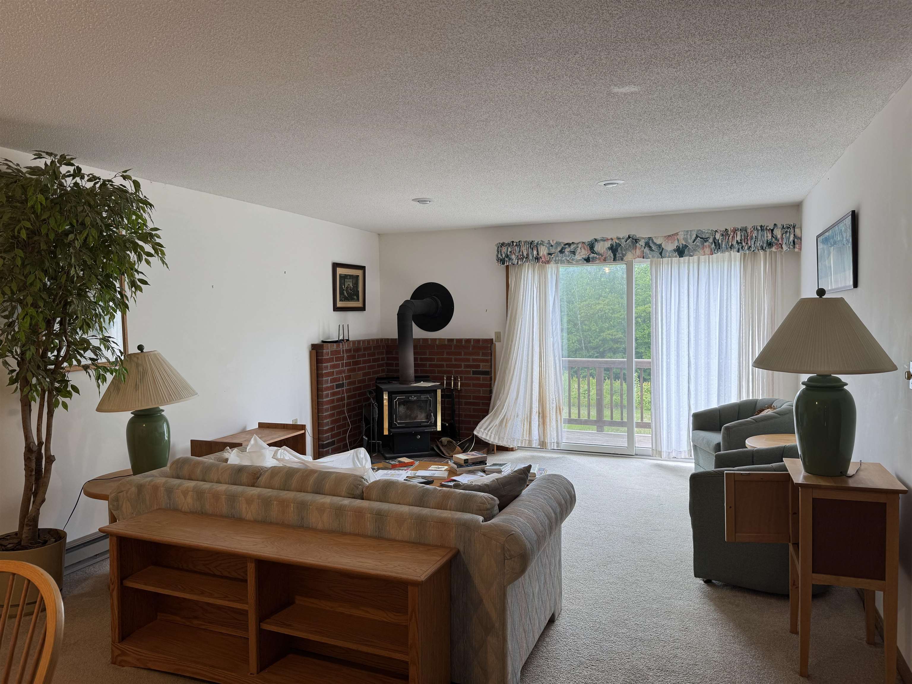 Furnished living room with a wood-burning stove, brick hearth, and sliding doors to a tree-lined balcony.