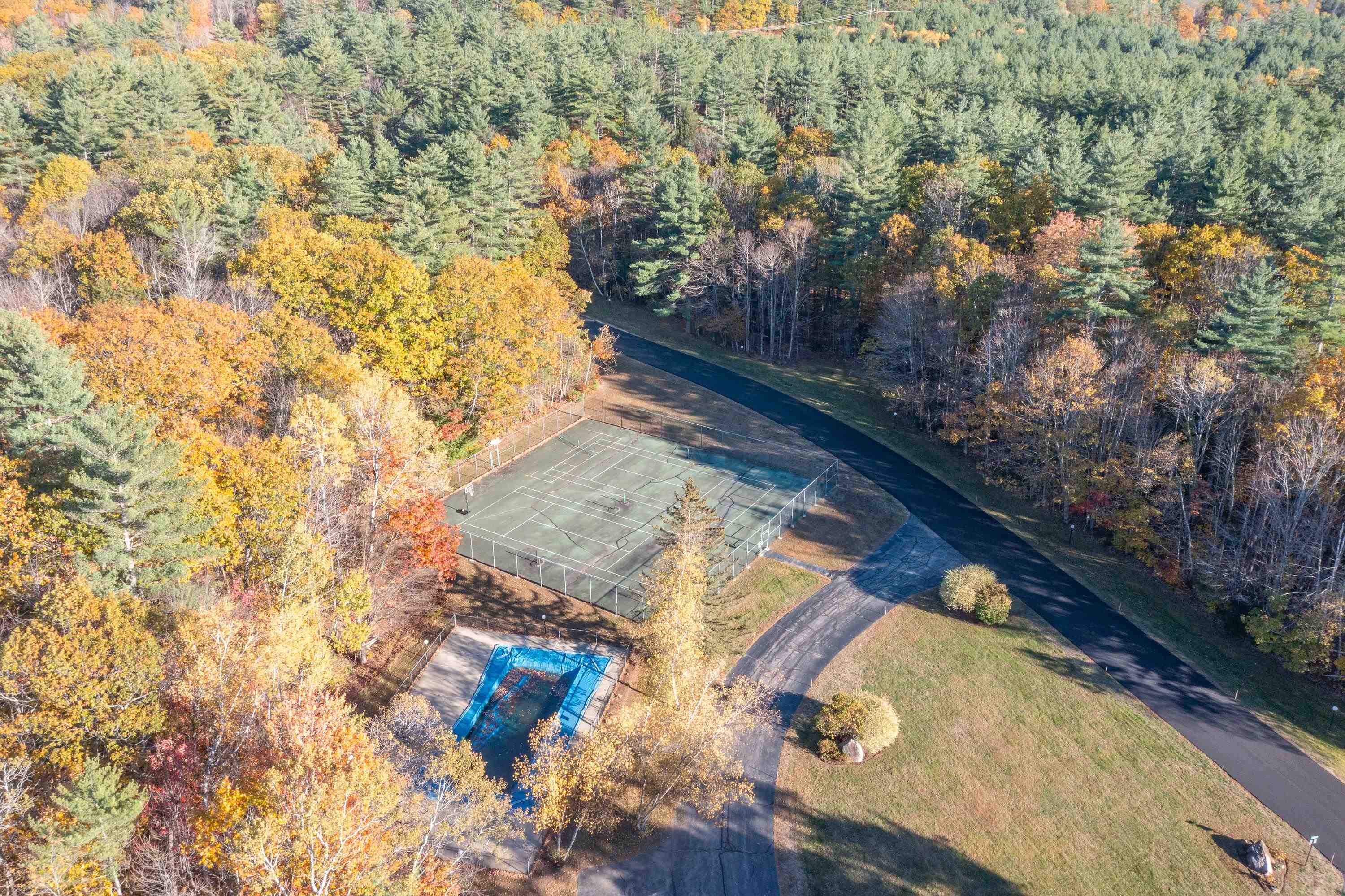 Aerial view of a tennis court and outdoor pool amid autumn woodland.