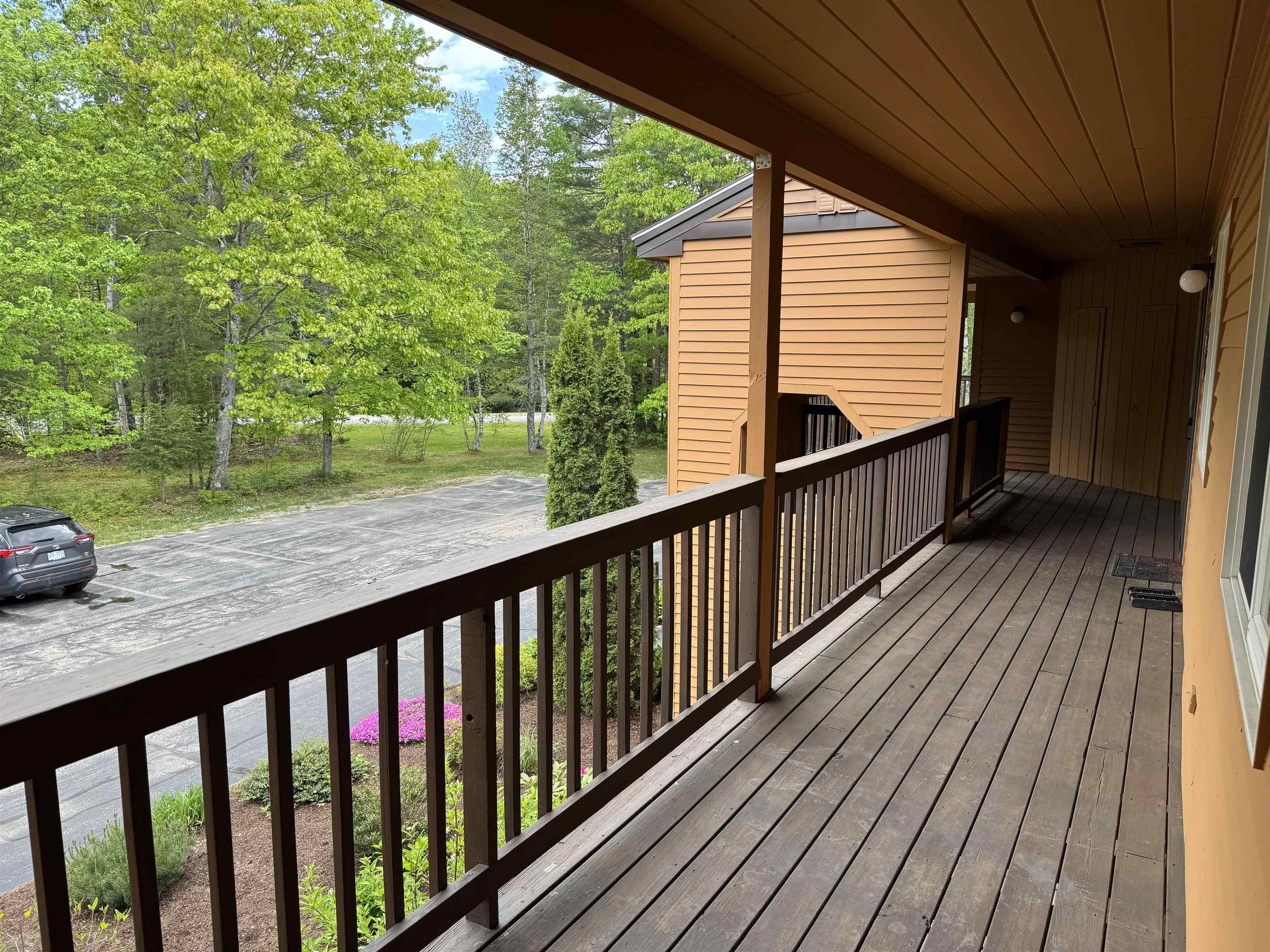Covered wooden porch overlooking landscaped grounds, a parking area, and leafy trees.