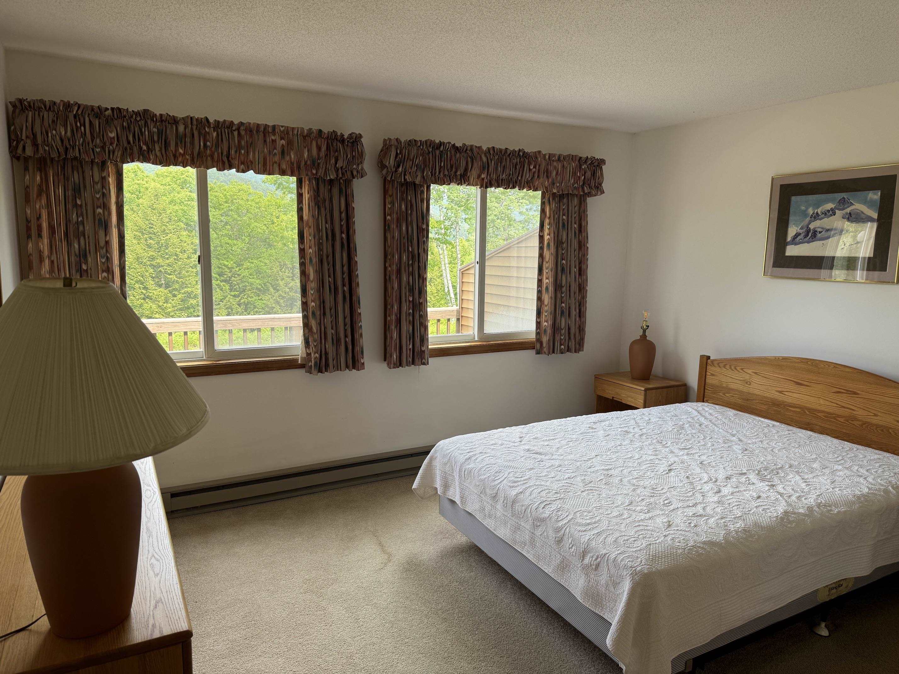 Bedroom with a white quilted bed and two curtained windows overlooking leafy trees.
