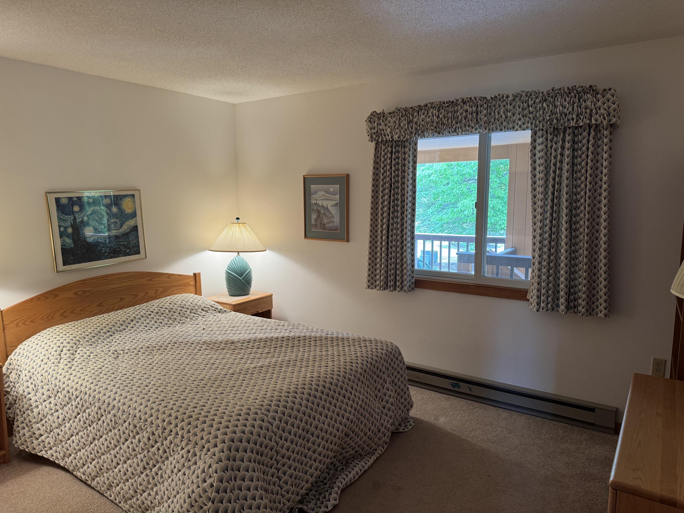Furnished bedroom with a patterned bedspread, wooden headboard, and window overlooking greenery.