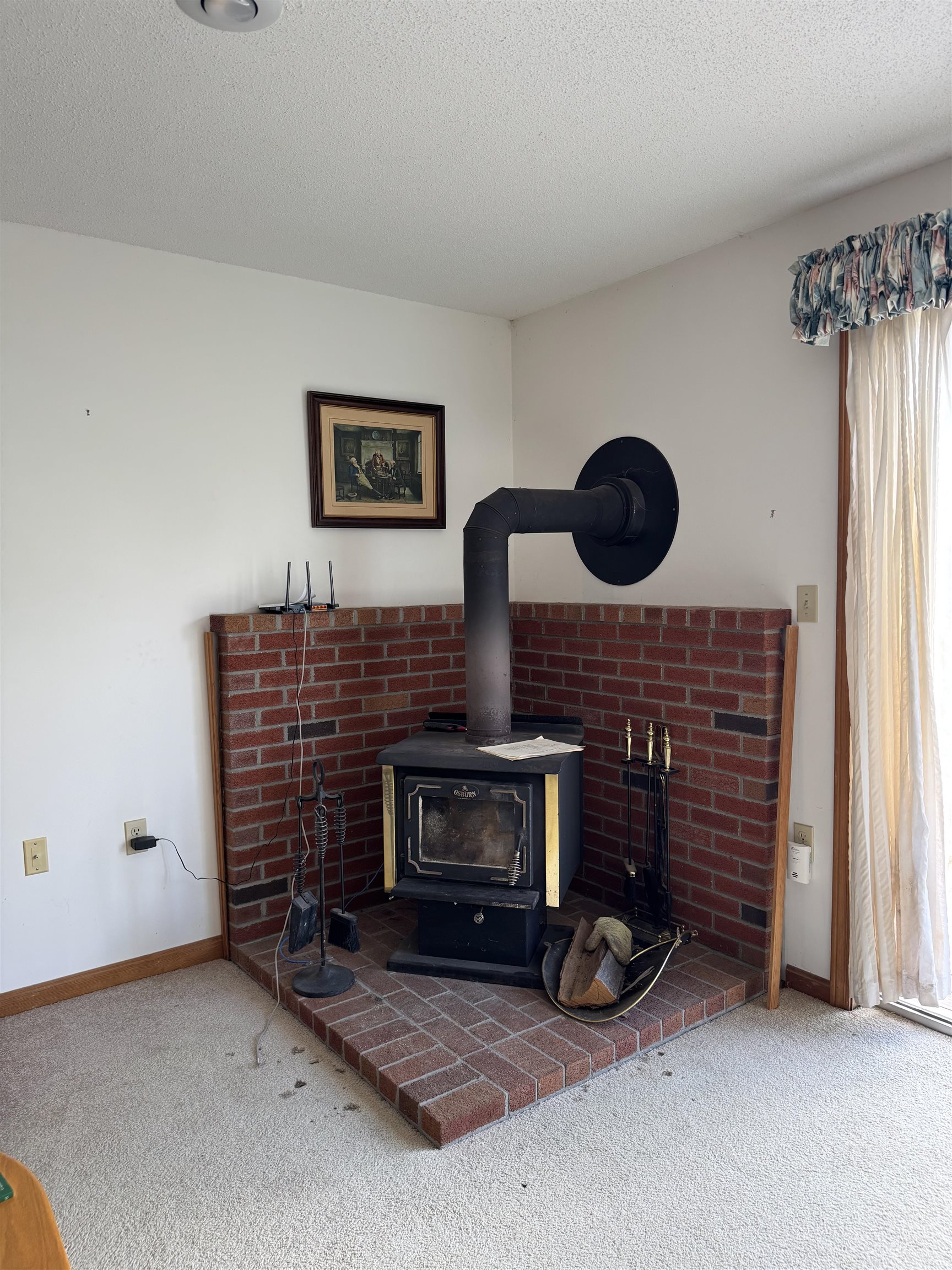 Living room wood stove set against a red brick hearth, beside a curtained window.