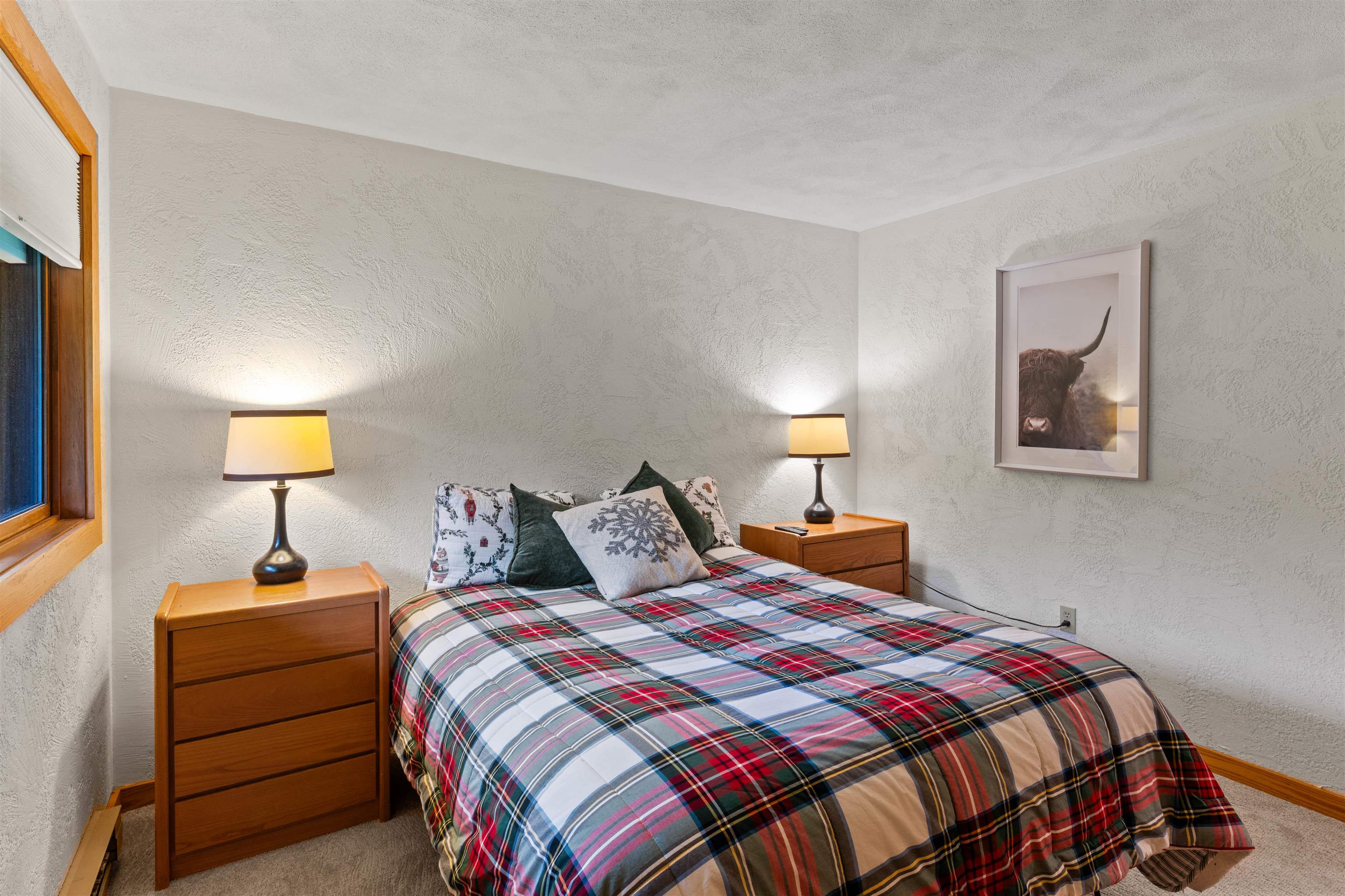 Bedroom with a plaid-covered bed, wooden nightstands, warm lamps, and a framed Highland cow print.