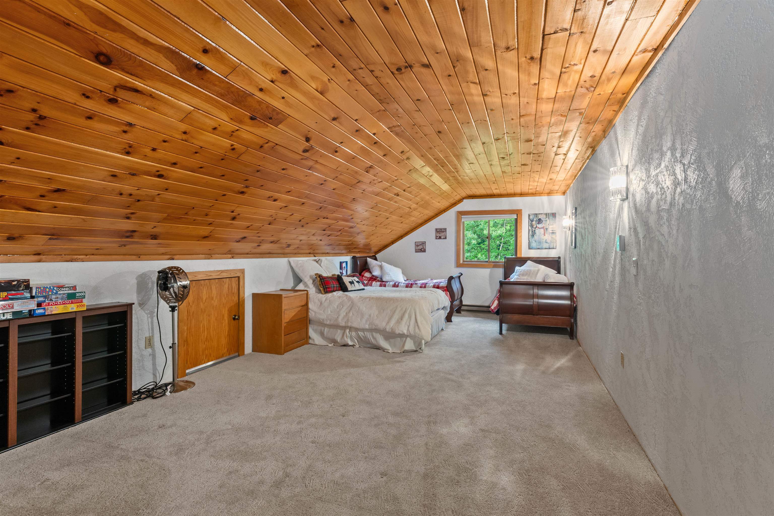 Attic bedroom with a sloped pine ceiling, multiple beds, and a window facing leafy trees.