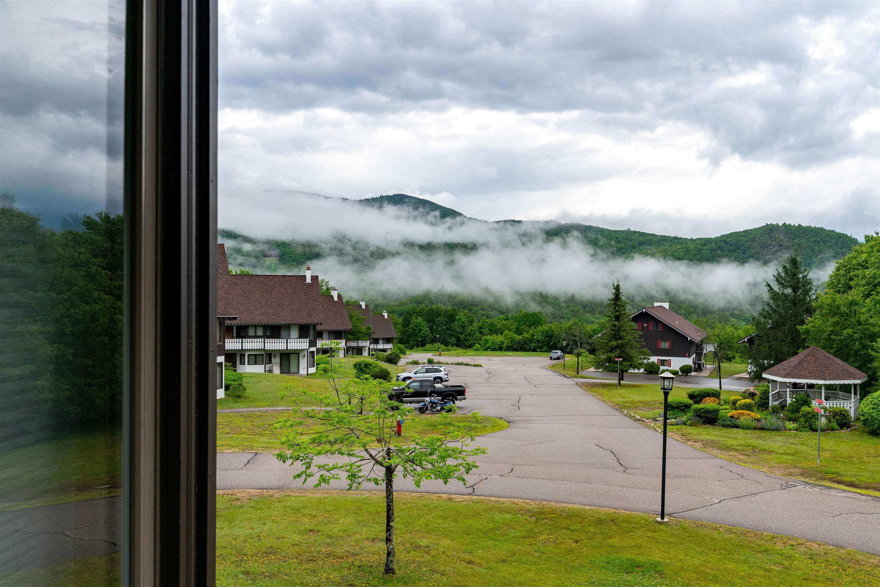 Mountain view over chalet-style buildings and a parking area, with low clouds drifting across green ridges.