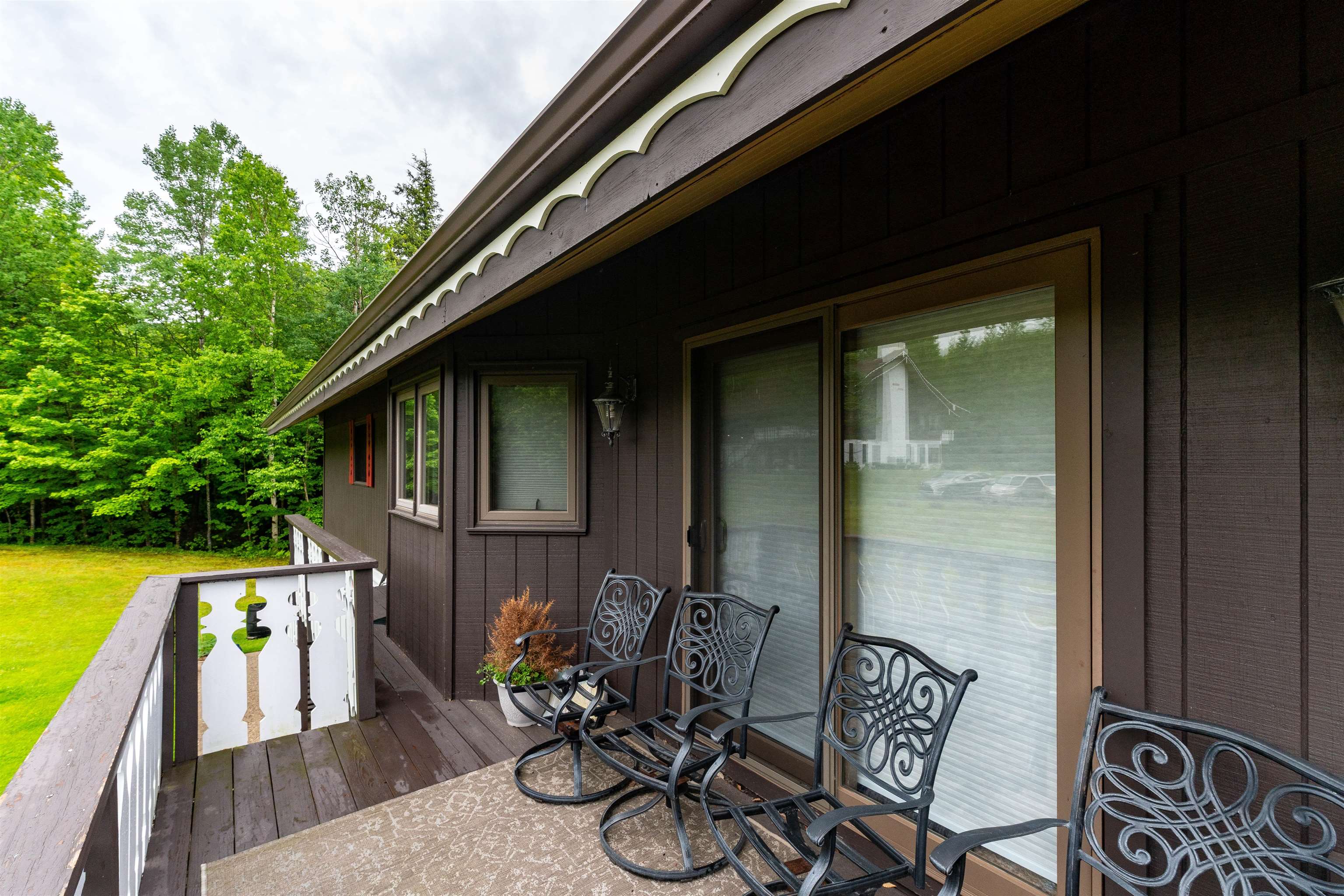 Wooden deck with ornate rocking chairs beside sliding glass doors, overlooking a lawn and trees.