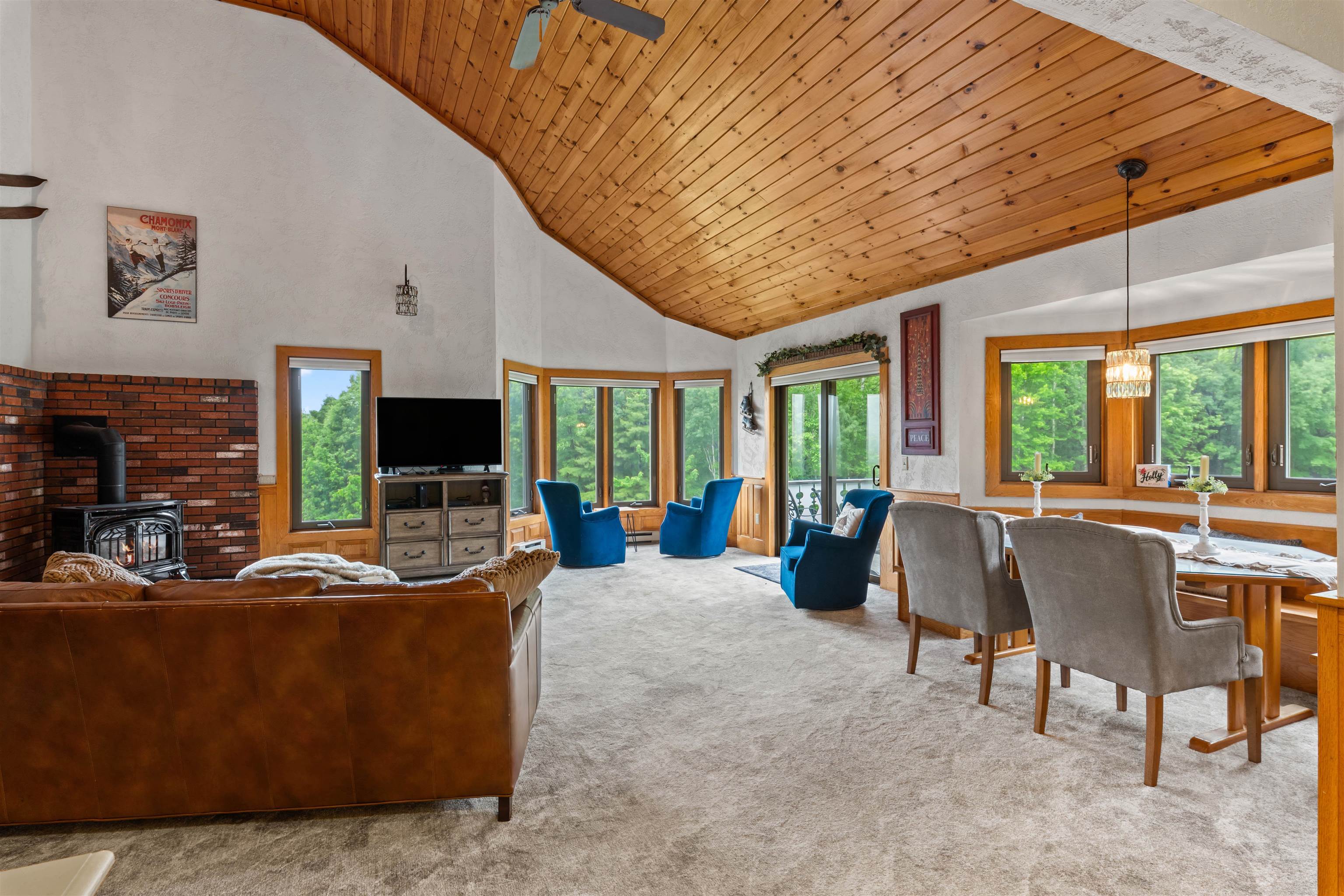 Living and dining room with vaulted wood ceiling, brick fireplace, and large windows overlooking trees.