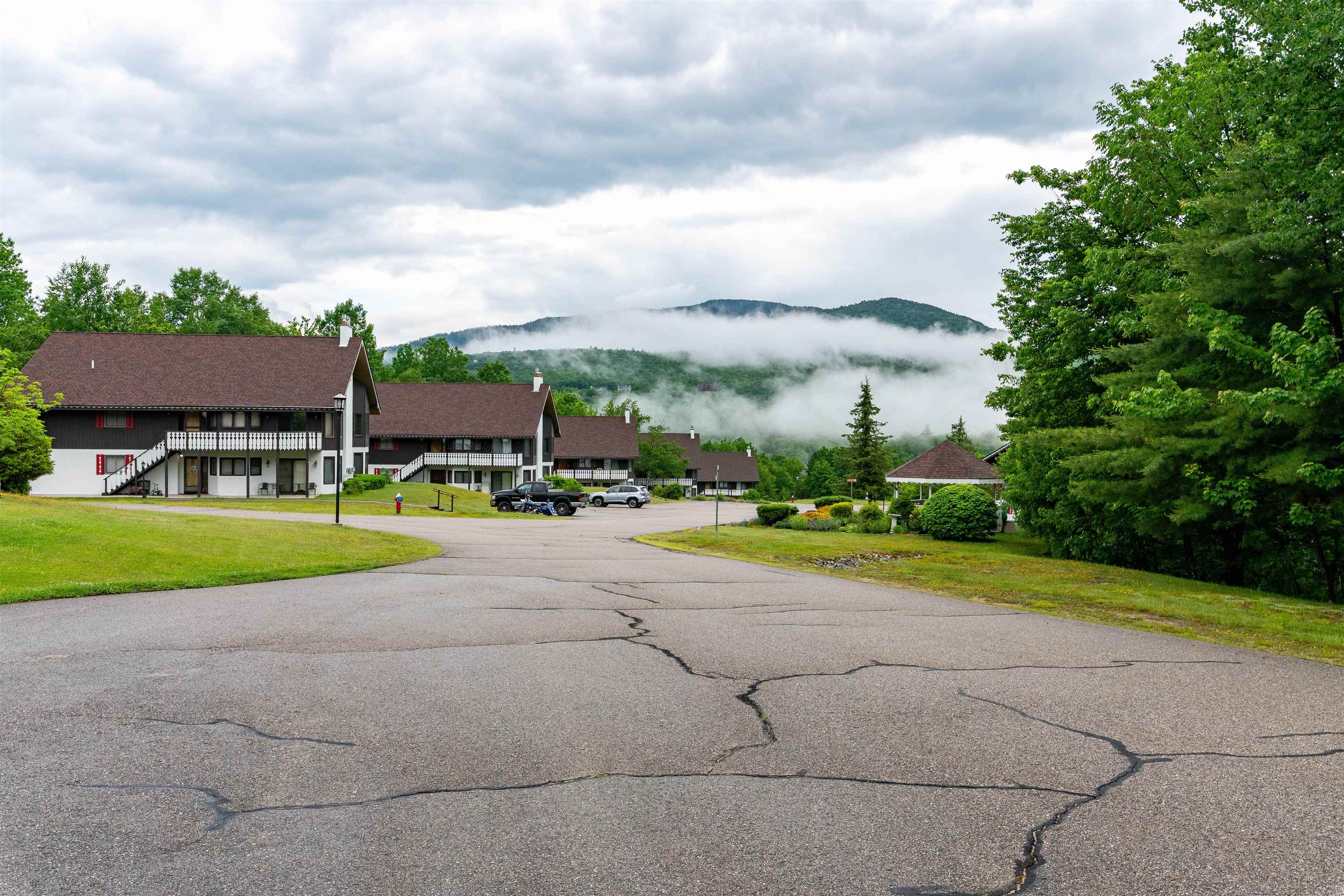 Chalet-style condominiums beside a paved drive, with a gazebo and misty mountain views.