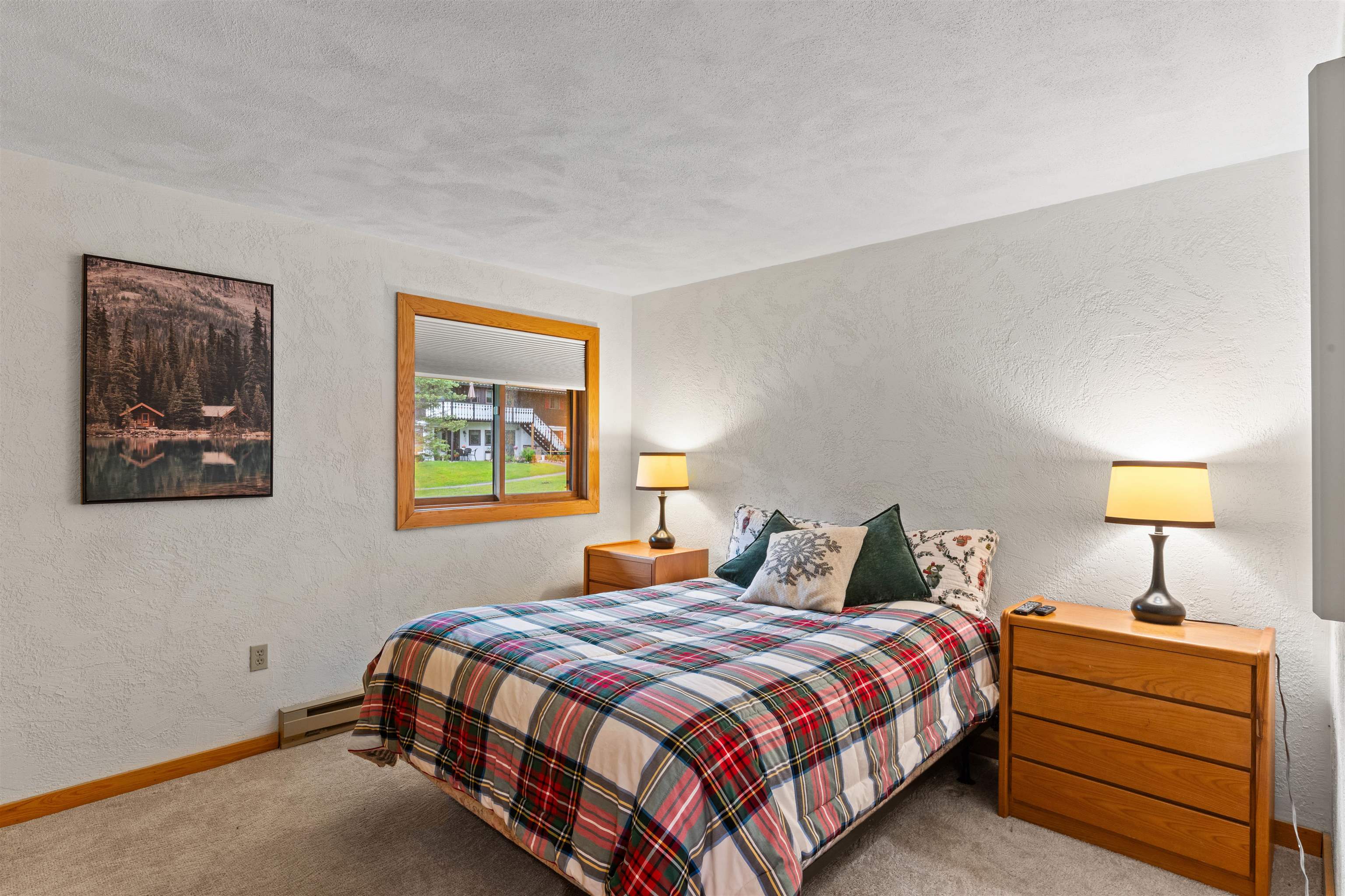 Furnished bedroom with a plaid-covered bed, wooden dresser, and window overlooking a courtyard.