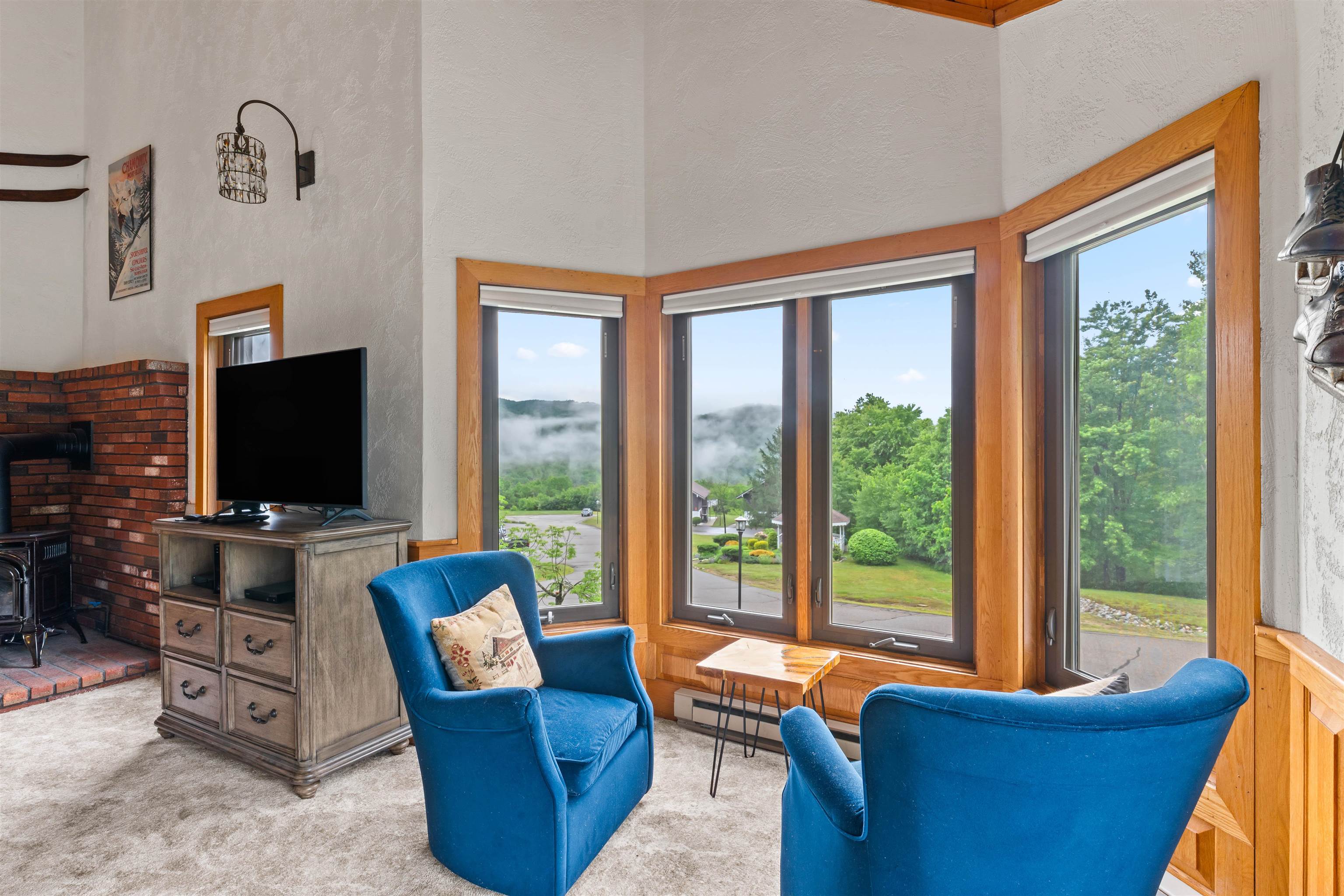 Living room with blue armchairs, a brick fireplace, and broad windows overlooking misty wooded hills.