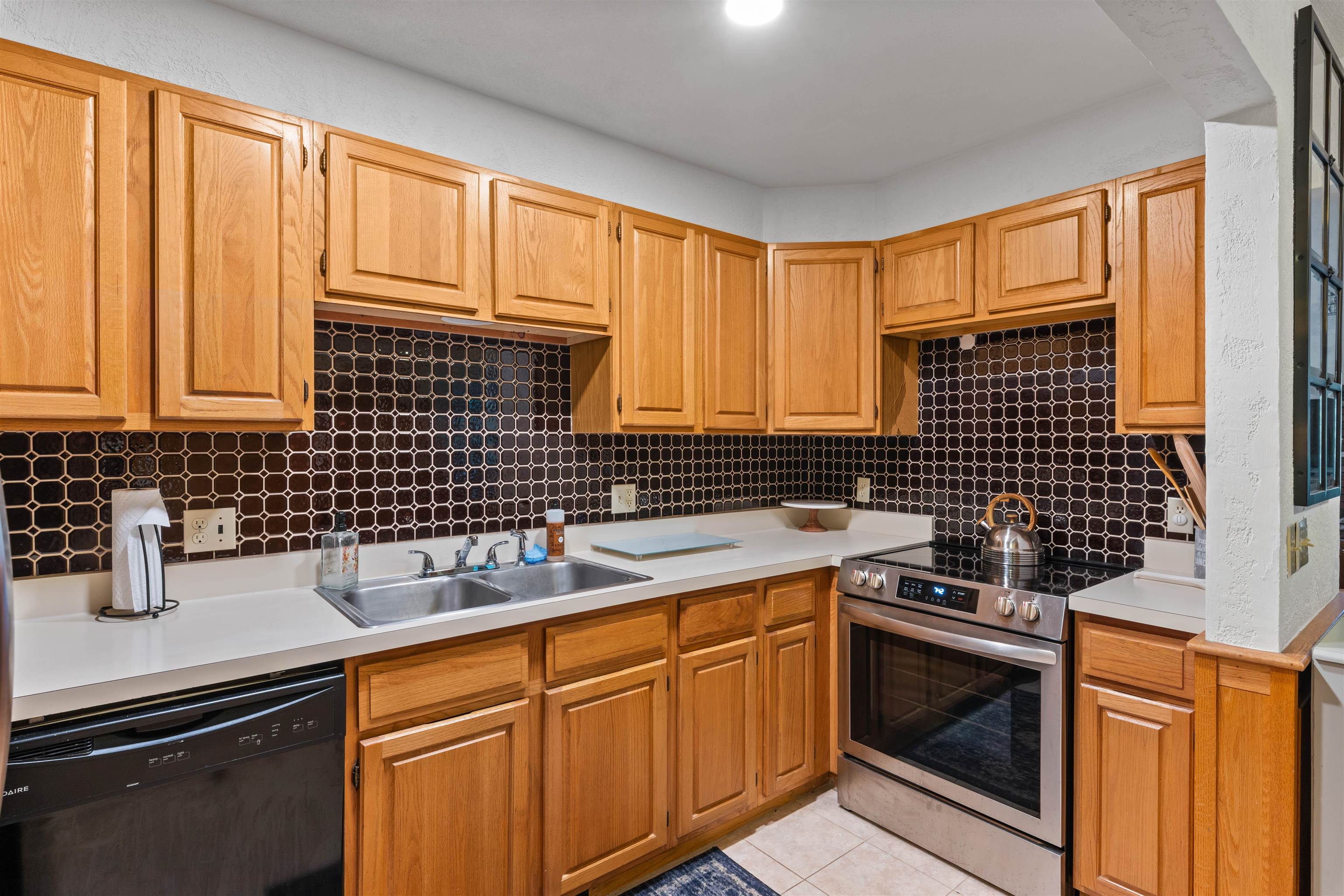 Kitchen with wood cabinets, dark geometric tile backsplash, white counters, stainless steel range and sink.