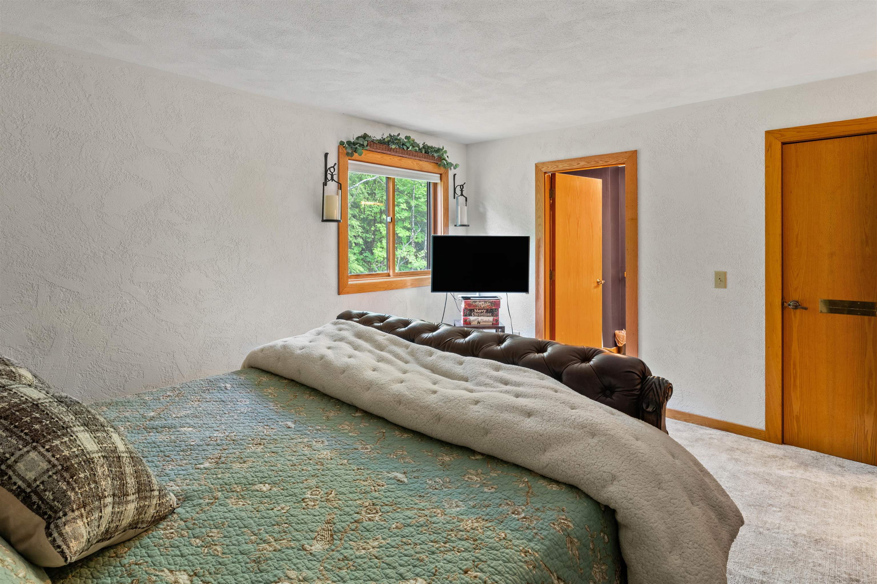 Bedroom with a green quilt, plush white blanket, dark tufted headboard, and wood-trimmed window.