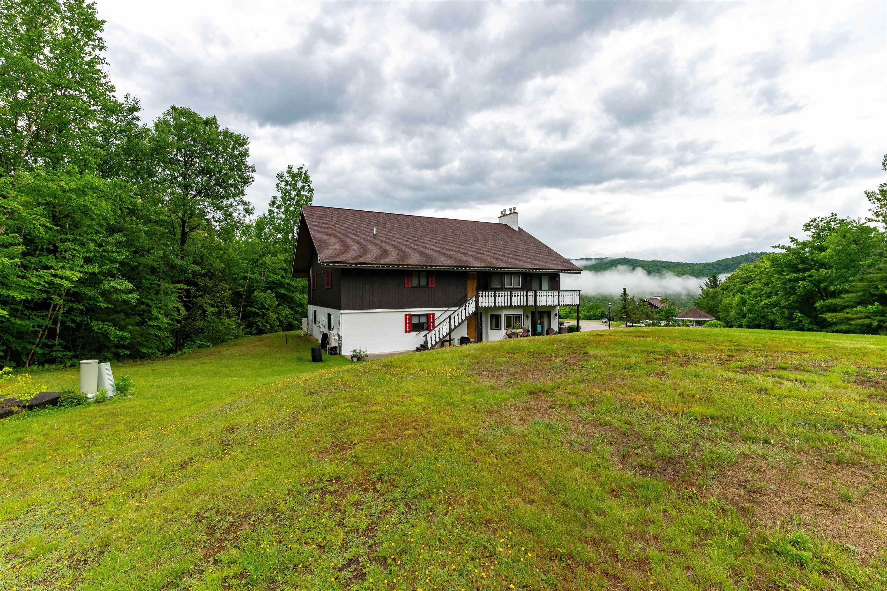 Two-story home with a balcony and exterior staircase above a sloping lawn, backed by misty forested hills.