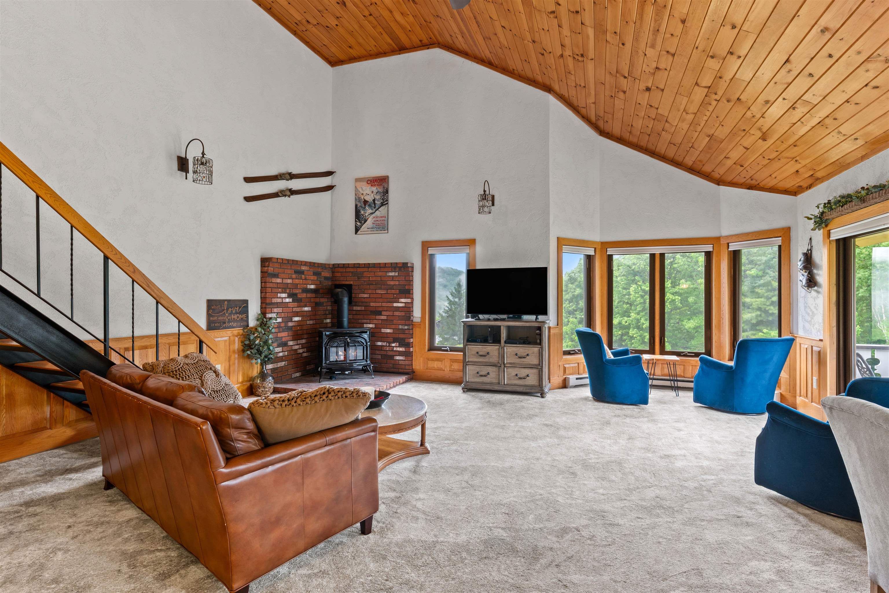 Living room with vaulted wood ceiling, brick fireplace, leather sofa, and blue chairs beside large windows.